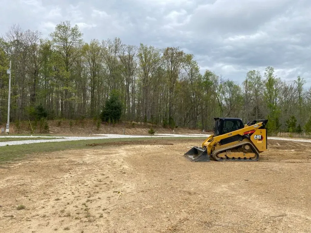 Yellow skid steer loader on a construction site, clearing land near trees and a cloudy sky.