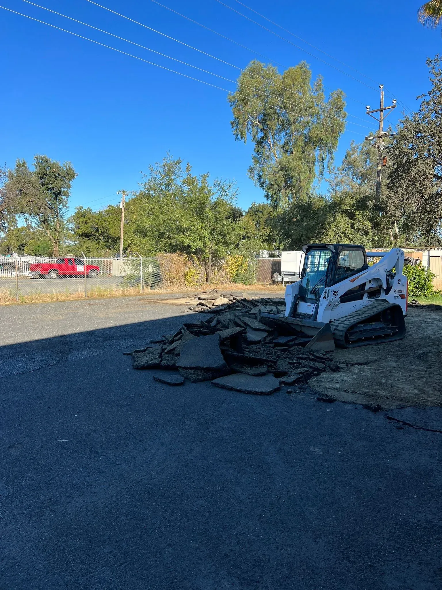 Bobcat removing asphalt on a sunny day. Dark gray pavement, bright blue sky, green trees.