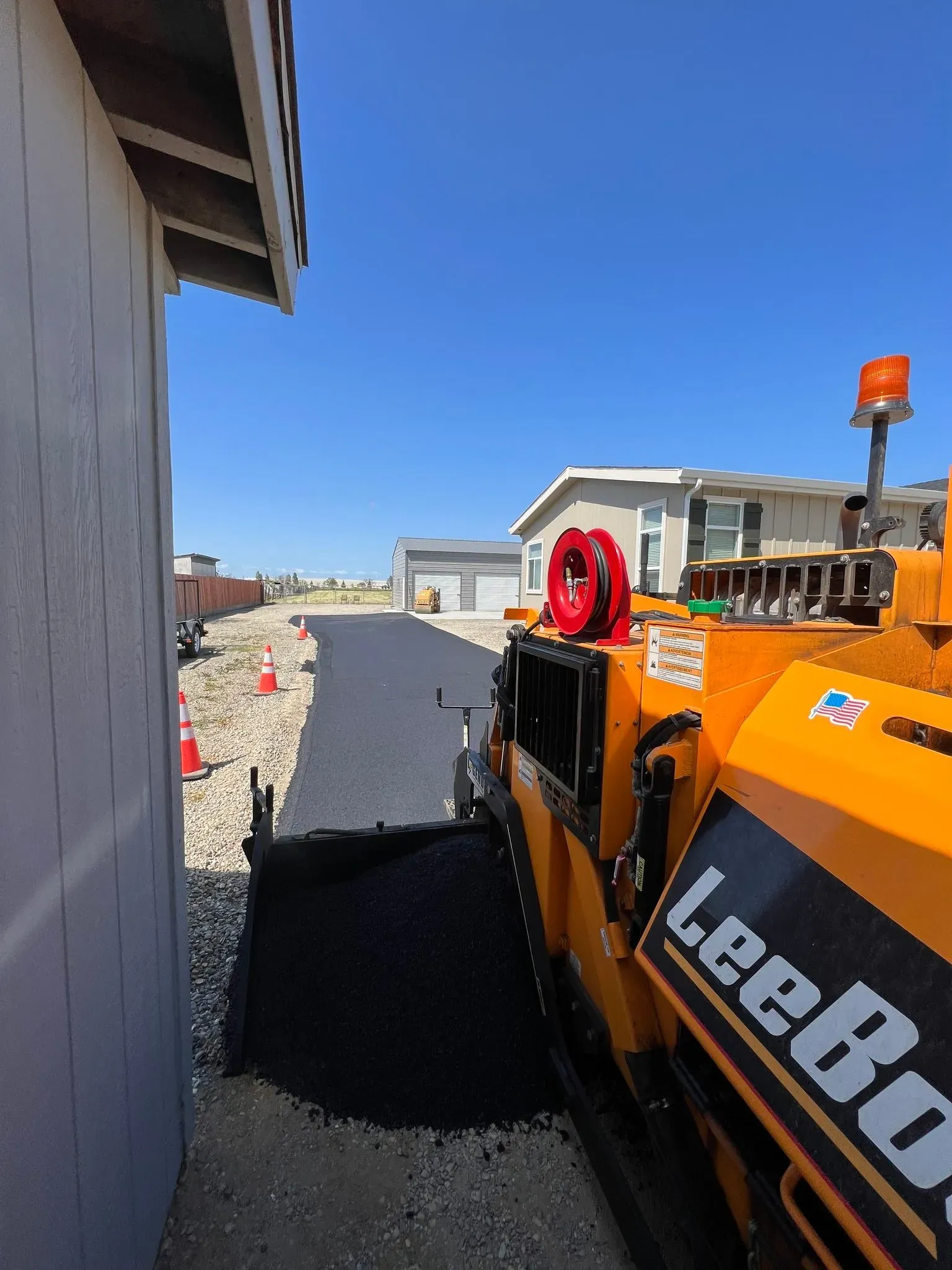 Asphalt paving machine laying asphalt on a driveway next to a building under a blue sky.