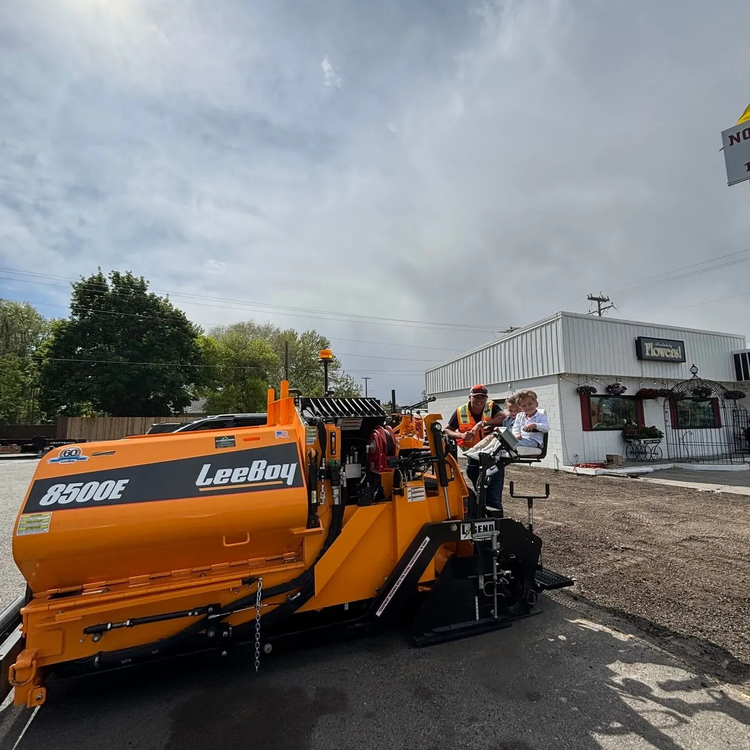 Orange road paving machine in front of a white building, under cloudy skies. Crew members visible.