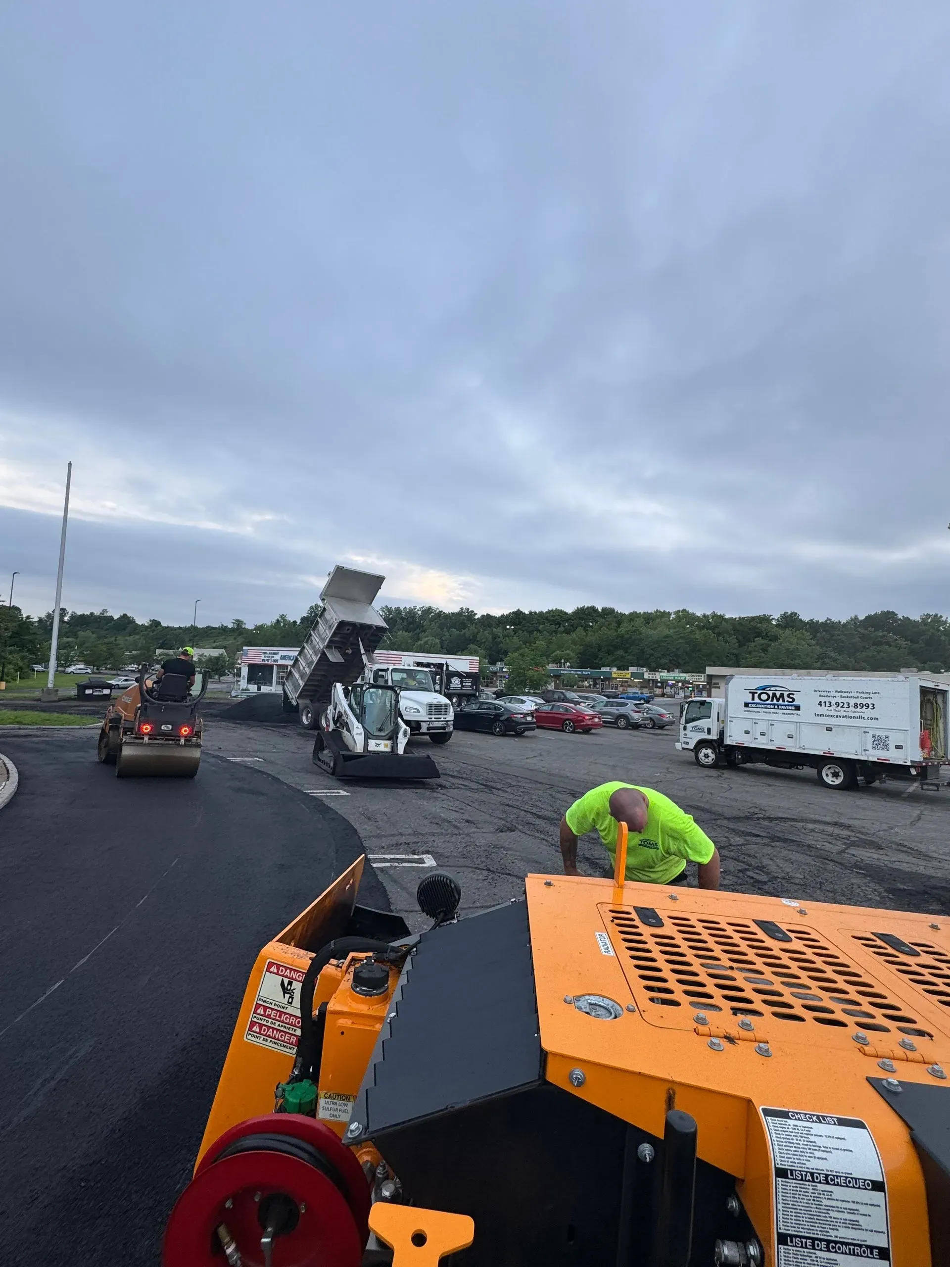 Asphalt paving in progress: worker in neon green, dump truck, and machinery on a cloudy day.