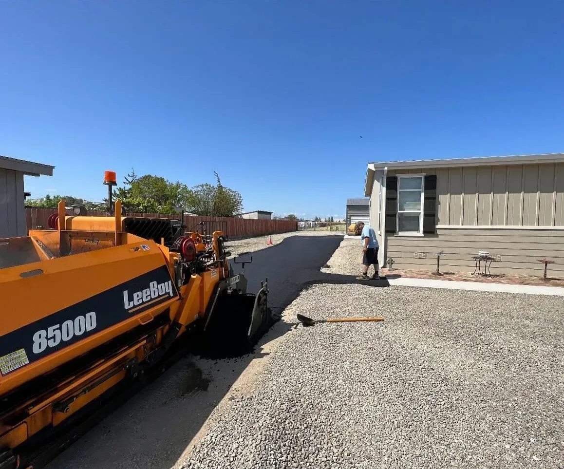 Asphalt paving machine laying a road in a residential area on a sunny day; a worker stands nearby.