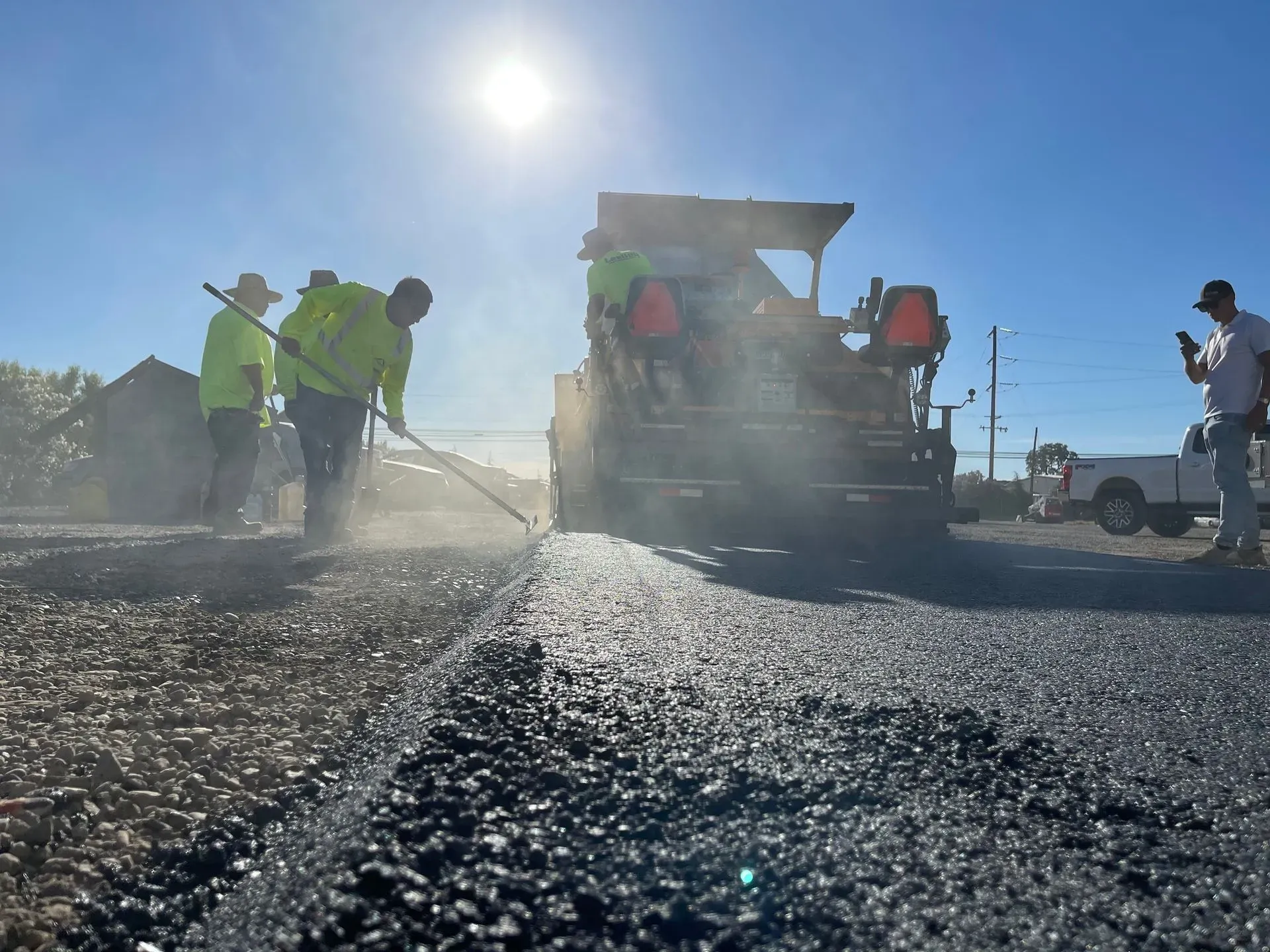 Road workers laying asphalt, one using a rake, near paving machine in bright sunlight.
