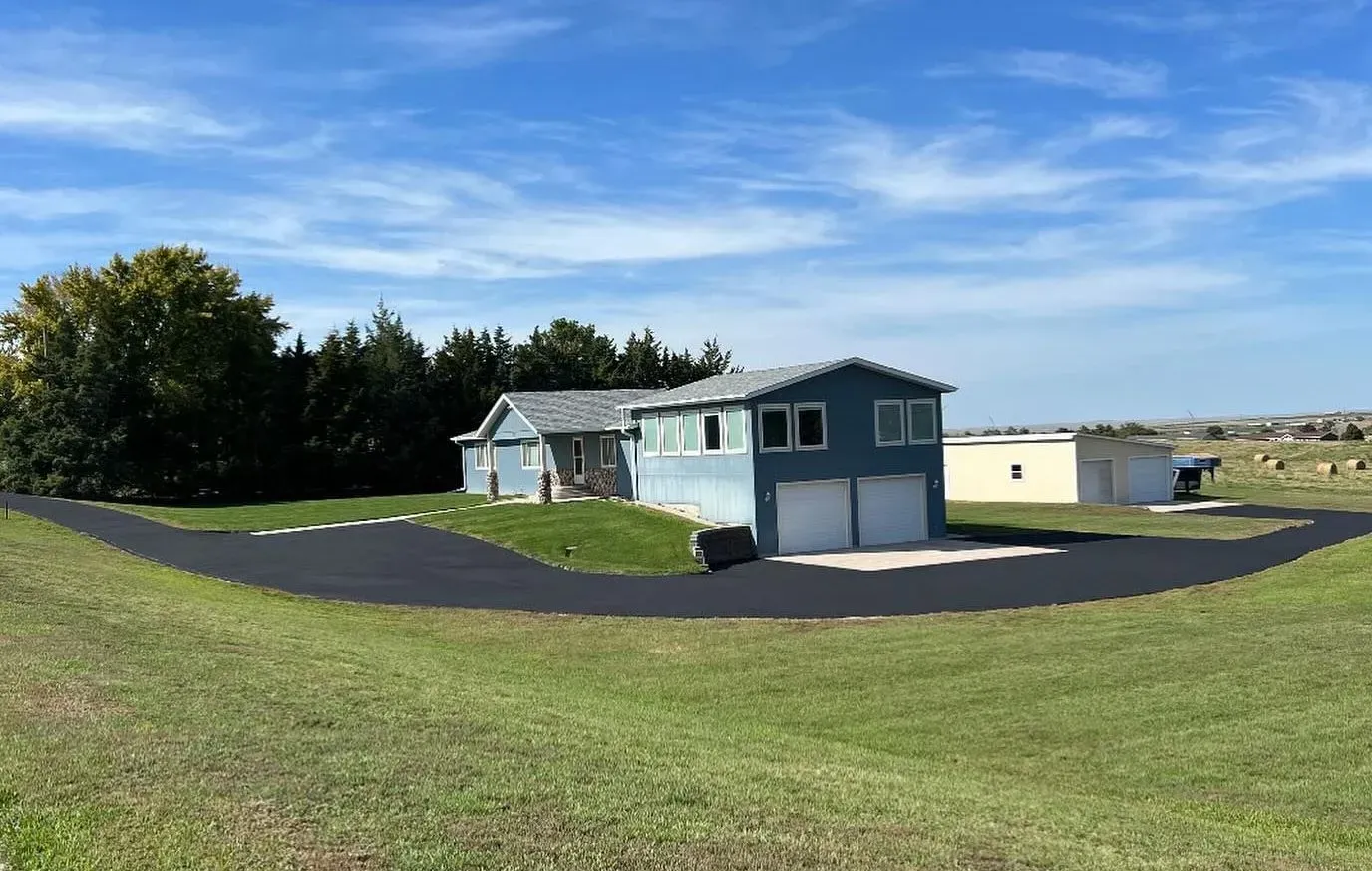 Blue house with a curved asphalt driveway, green lawn, and trees under a blue sky.