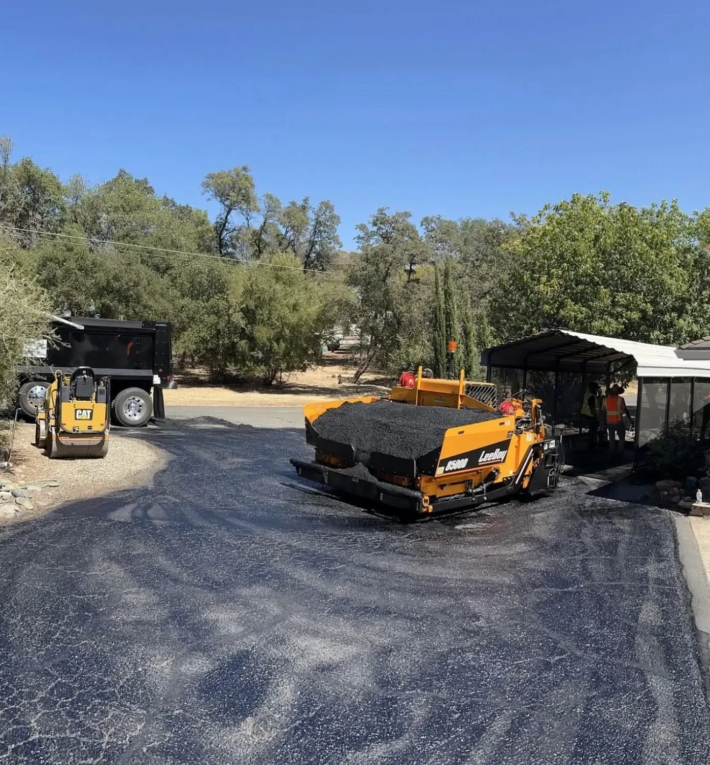 Asphalt paving in progress: yellow paver, dump truck, small loader, laying black asphalt on road.