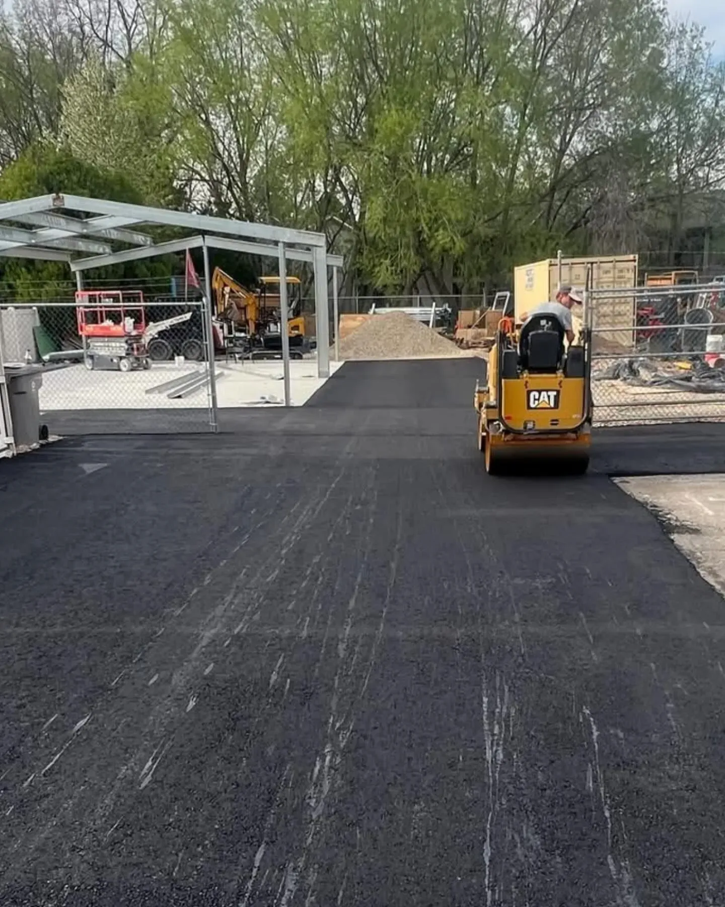 A worker compacts fresh asphalt with a roller in an outdoor construction zone, under a metal awning.