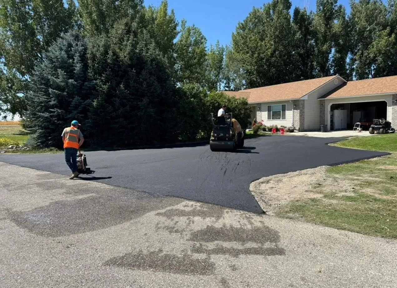 Workers paving a driveway in front of a house, with a roller and edger on a sunny day.