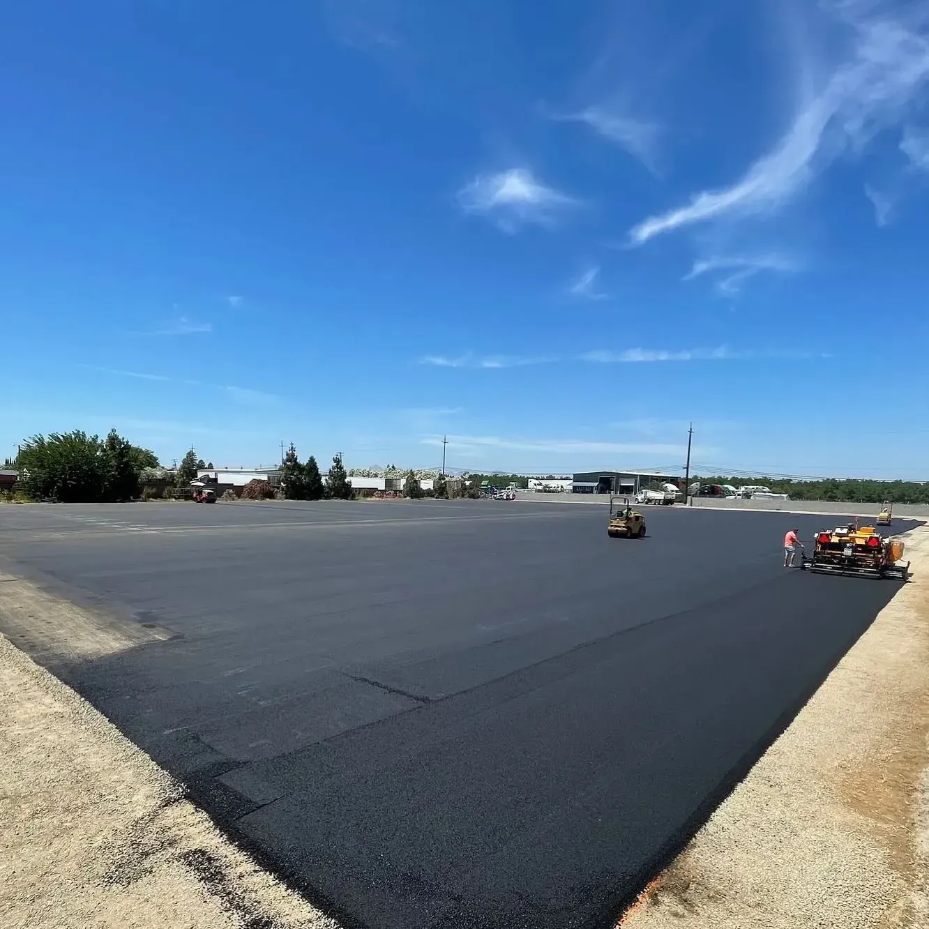 Asphalt paving in progress; machines laying black asphalt on a large, flat surface under a blue sky.