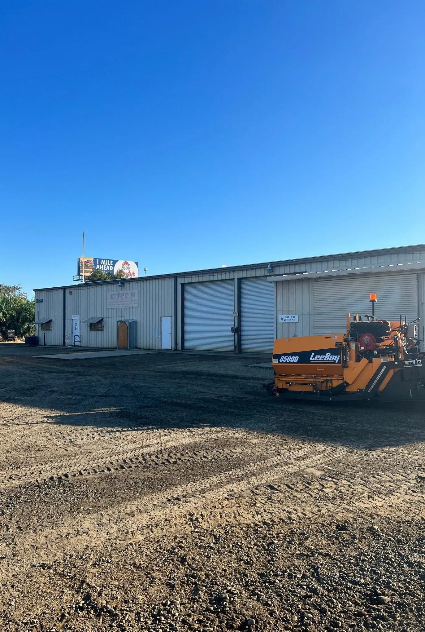 An orange paving machine in front of a gray building with a clear blue sky above.