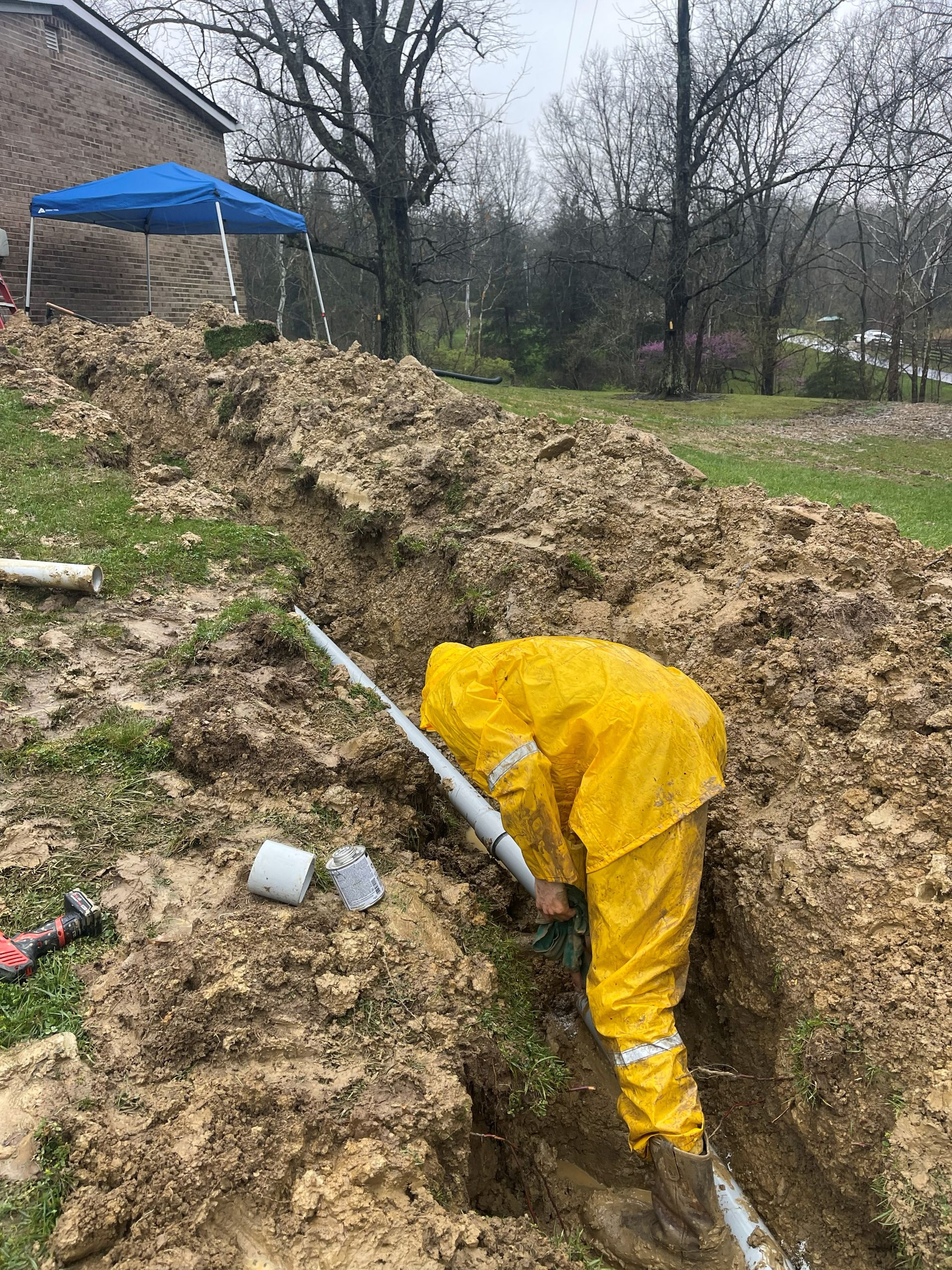 Person in yellow protective suit working on a pipe in a muddy trench outdoors.
