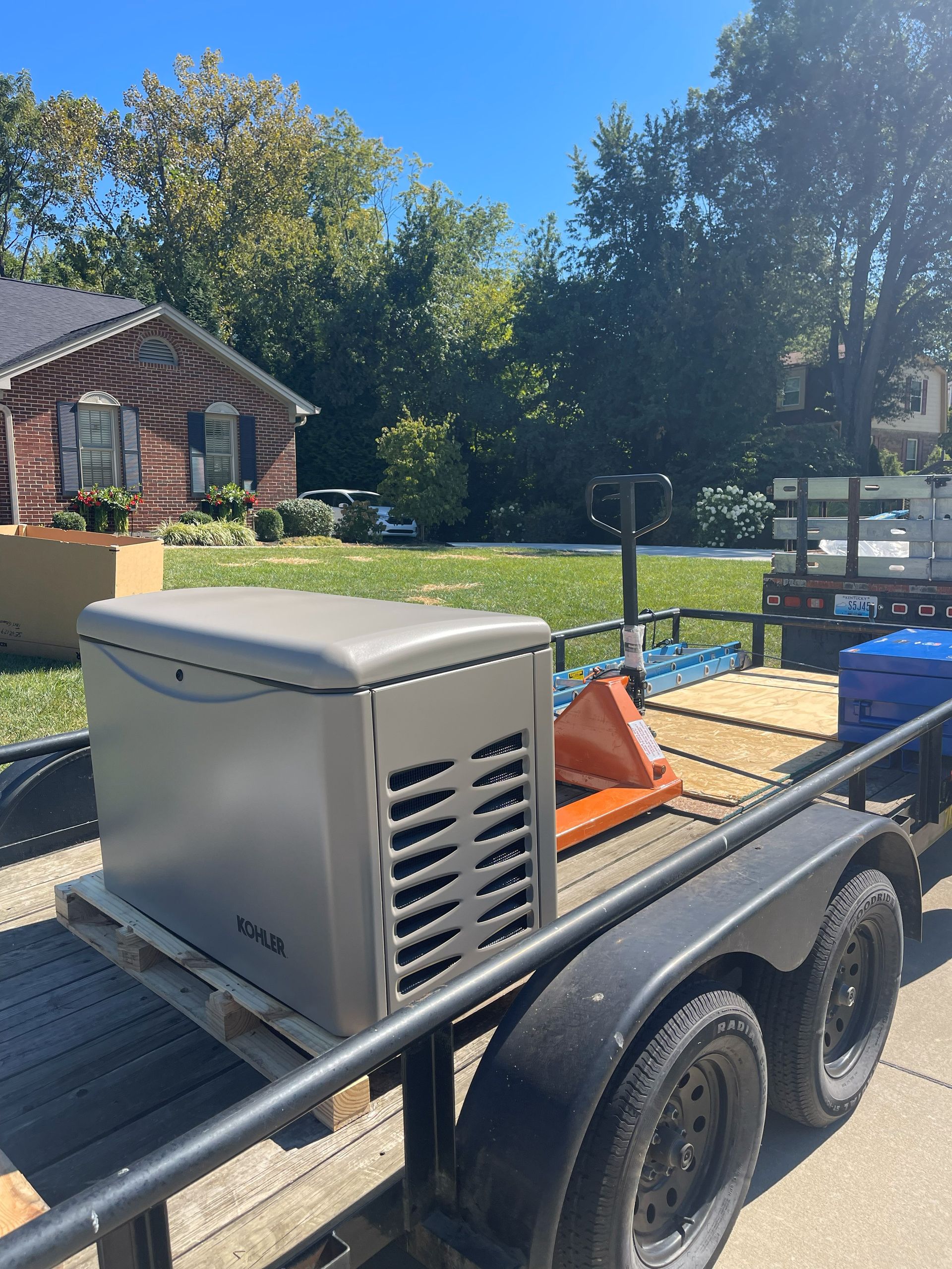 Generator on a trailer in front of a house. The generator is gray. Blue skies and trees are in the background.