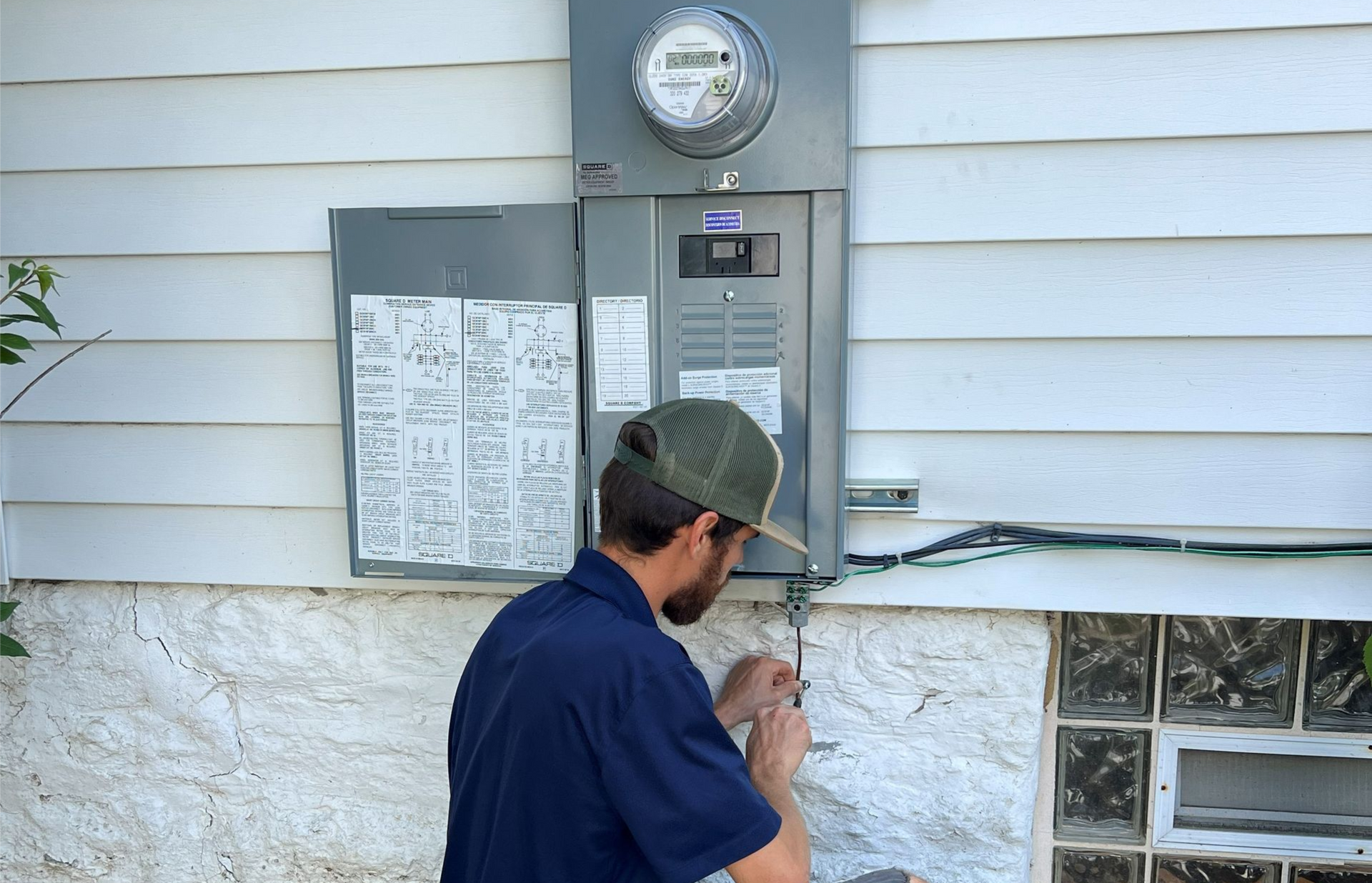 Man crouches, working on electrical box on the side of a house.