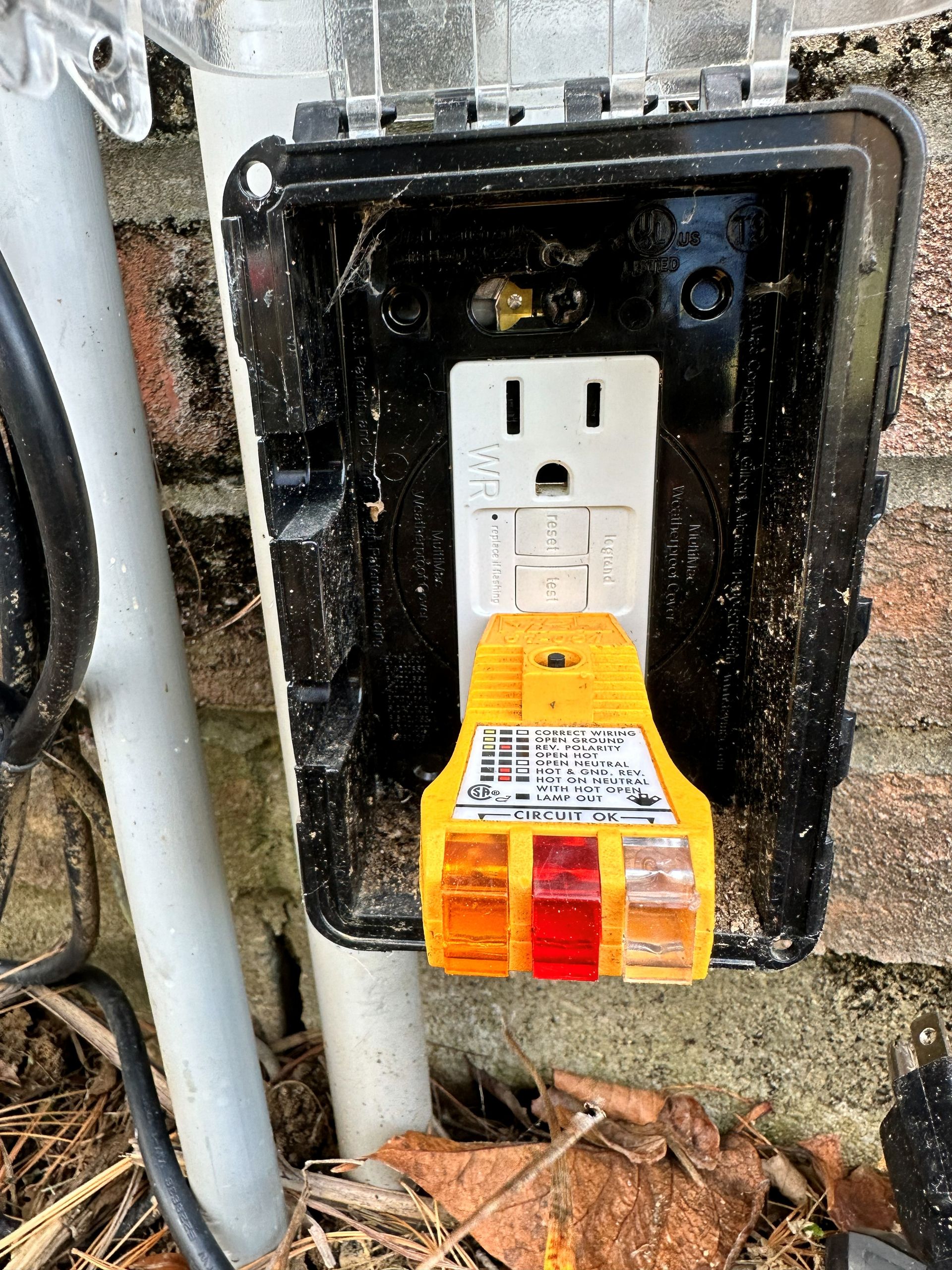 Outdoor electrical outlet with a yellow circuit tester, set in a black weatherproof box, next to a pipe.