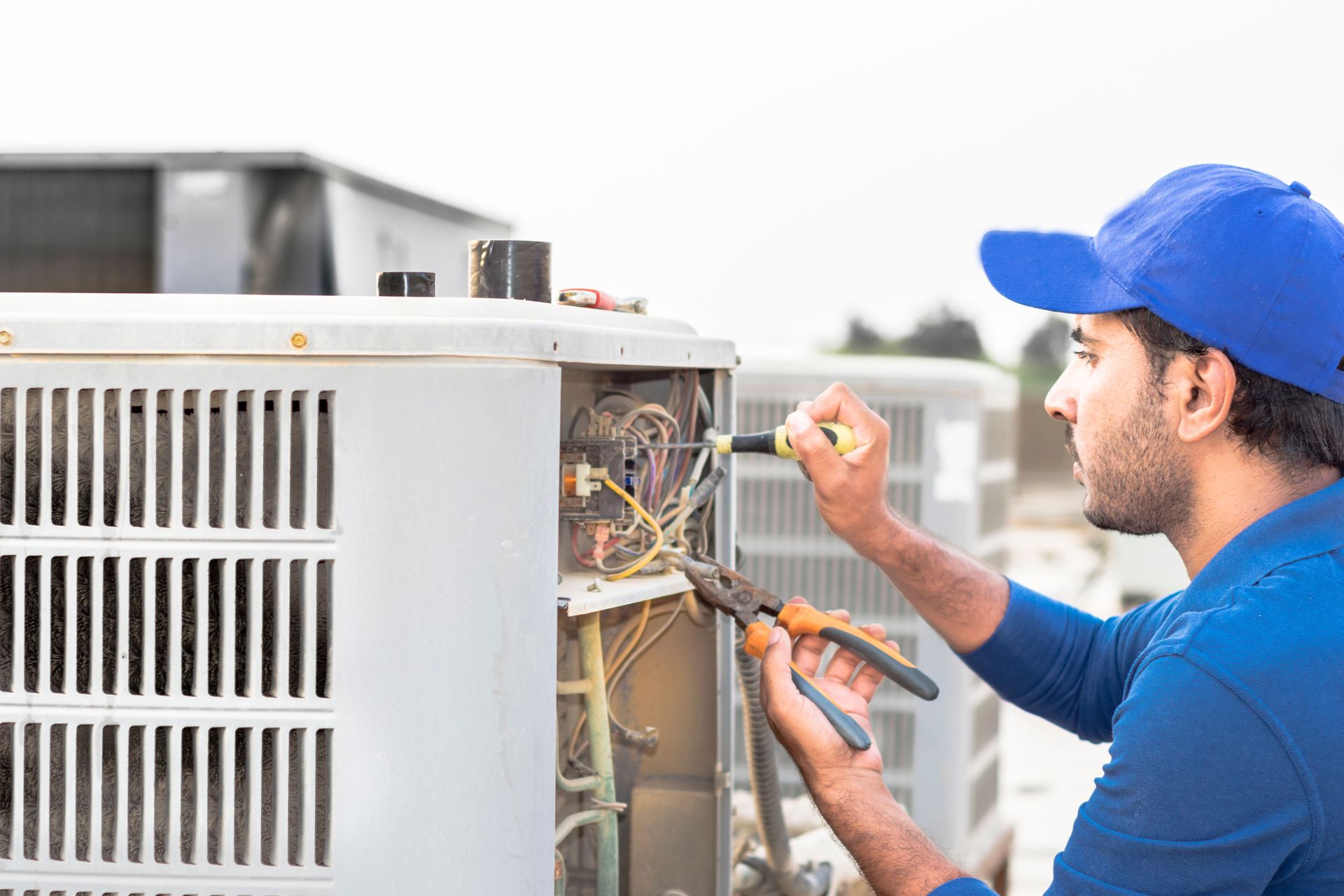 HVAC technician in blue cap repairs AC unit outdoors, using pliers and screwdriver.
