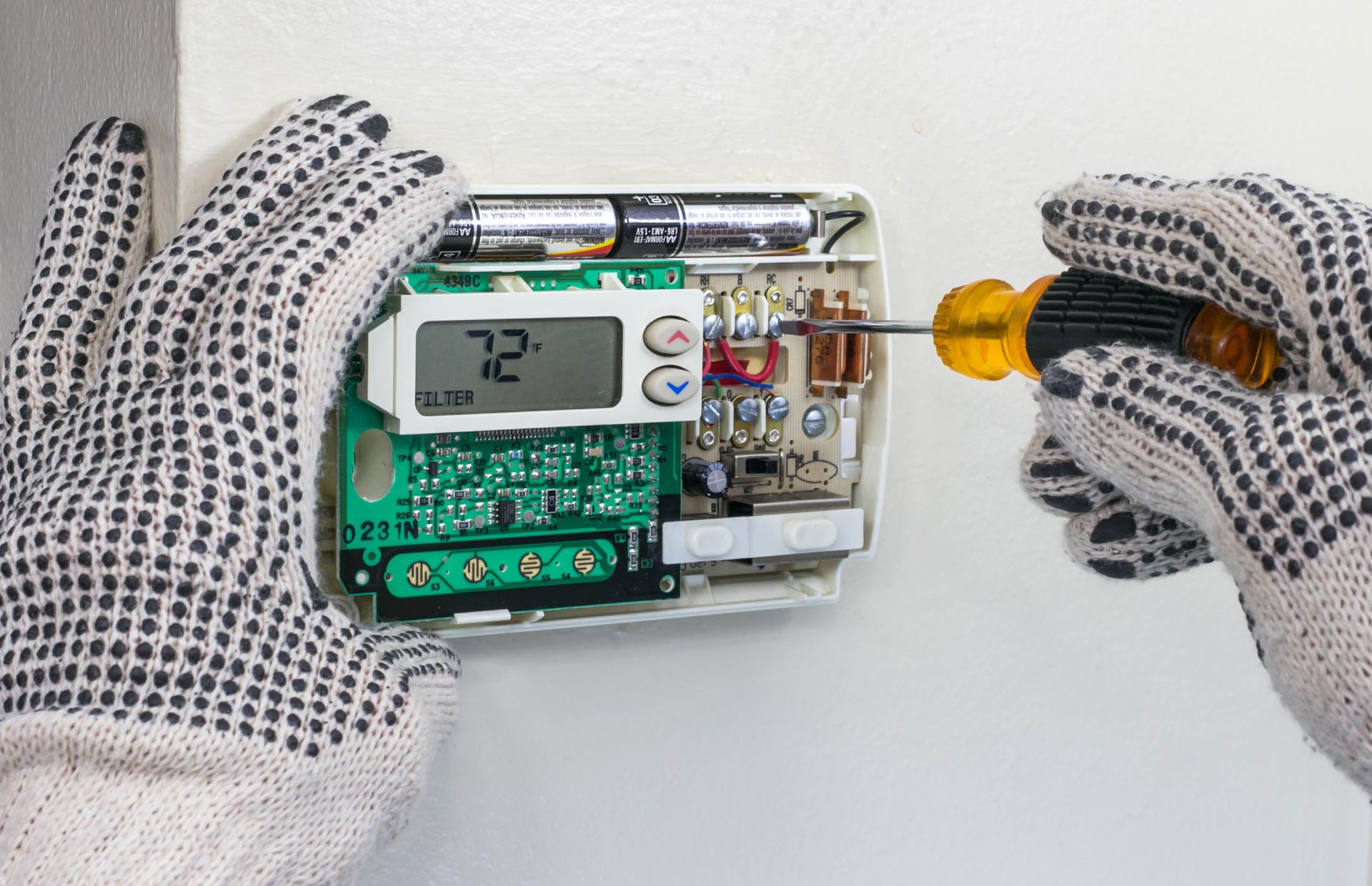 Person in gloves using screwdriver to work on a thermostat, set to 72 degrees, mounted on a wall.