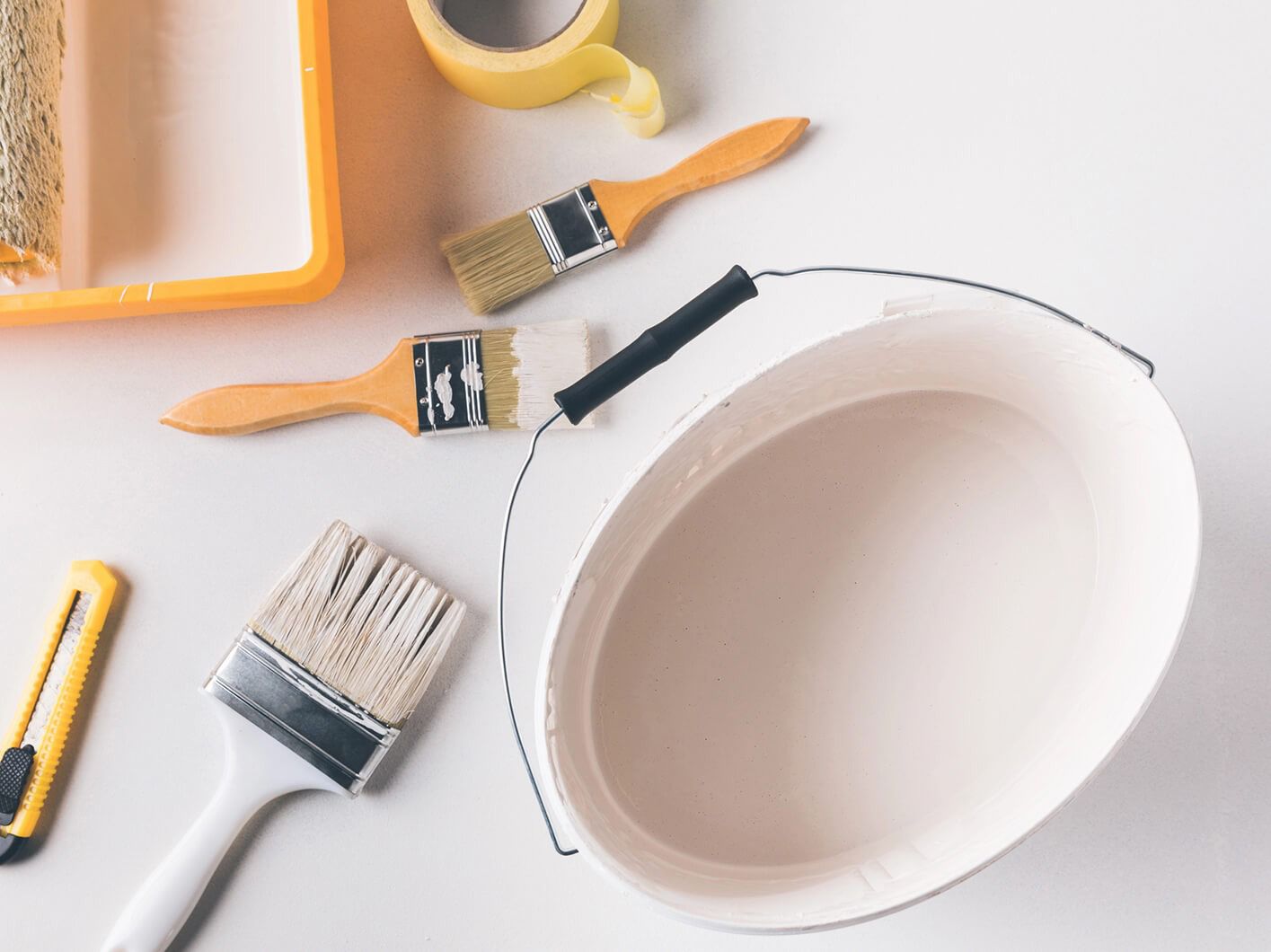 Painting supplies: paint bucket, brushes, roller tray, tape, utility knife on white surface.