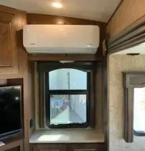 Air conditioner mounted above a window in a camper, with wood paneling and cabinetry.