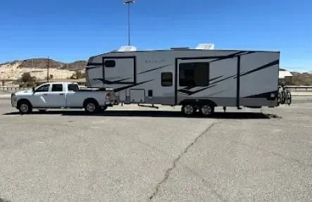 White pickup truck towing a large, gray and black travel trailer in a parking lot on a sunny day.
