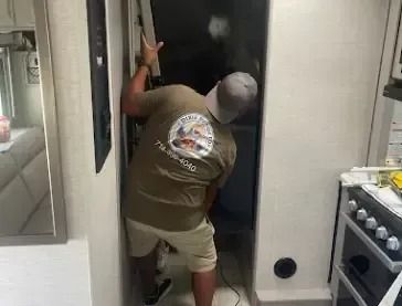 Person in a RV doorway, wearing a hat, cutting with a saw. White walls, gas range visible.