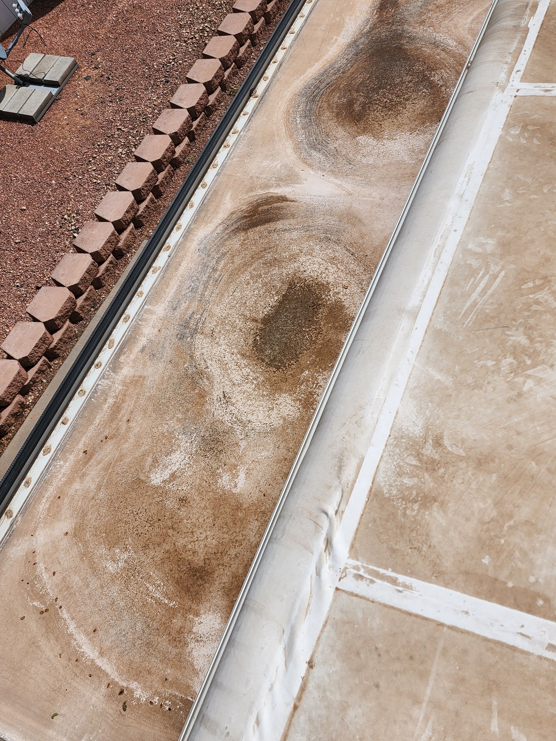 Overhead view of a flat roof with dirt and discoloration, possibly from standing water, near a brick edge.
