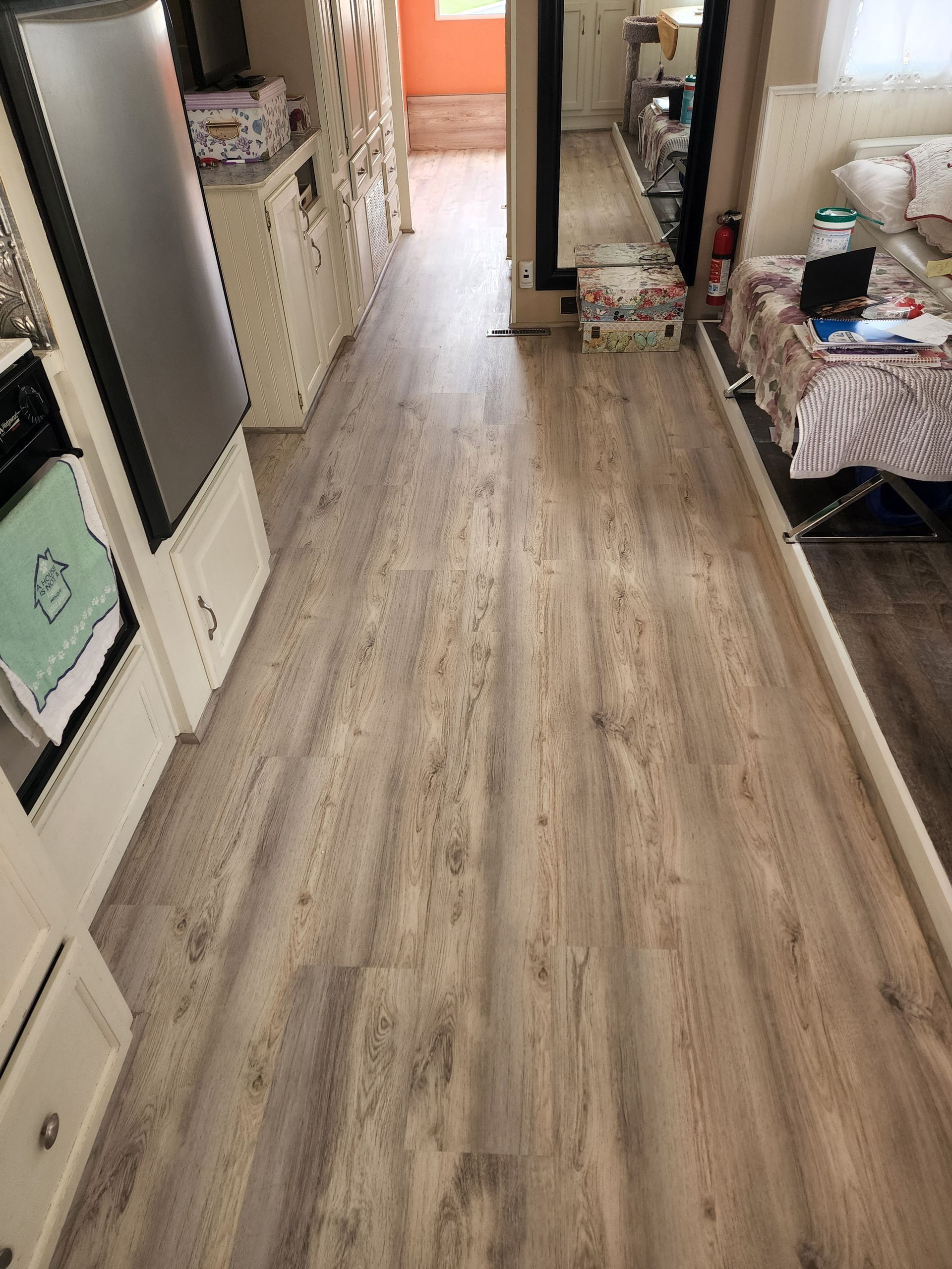 Interior view of a narrow RV hallway with light wood-look flooring. A refrigerator and cabinets line the left side.