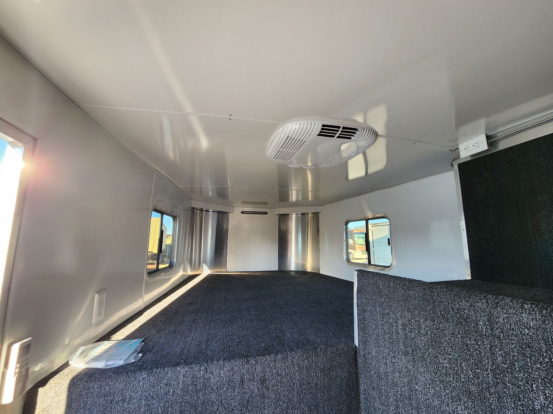 Interior of a horse trailer with a bed, windows, and ceiling fan. White walls, gray carpet.