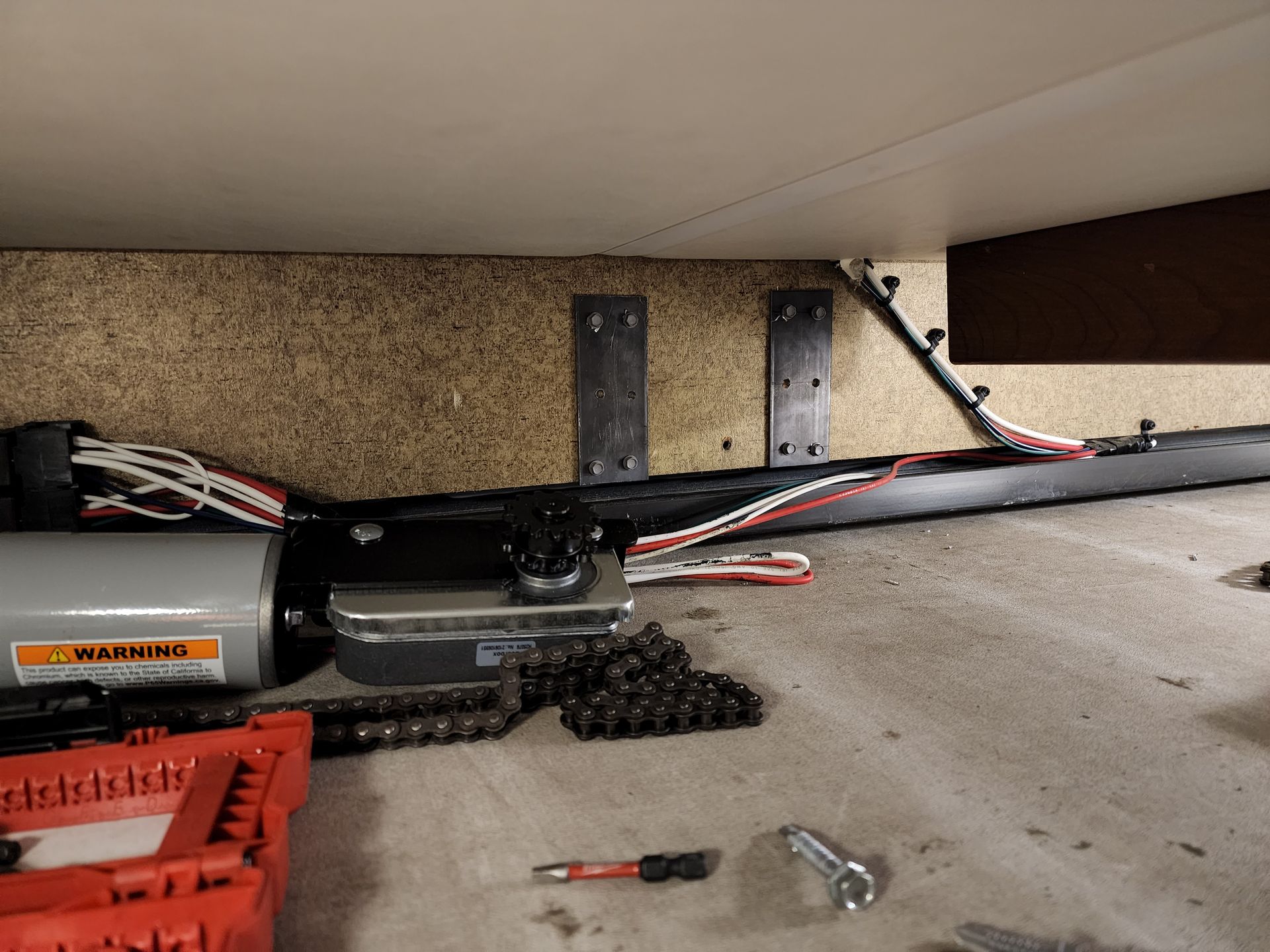 Garage door opener components and chain on a concrete floor beneath a wooden shelf.