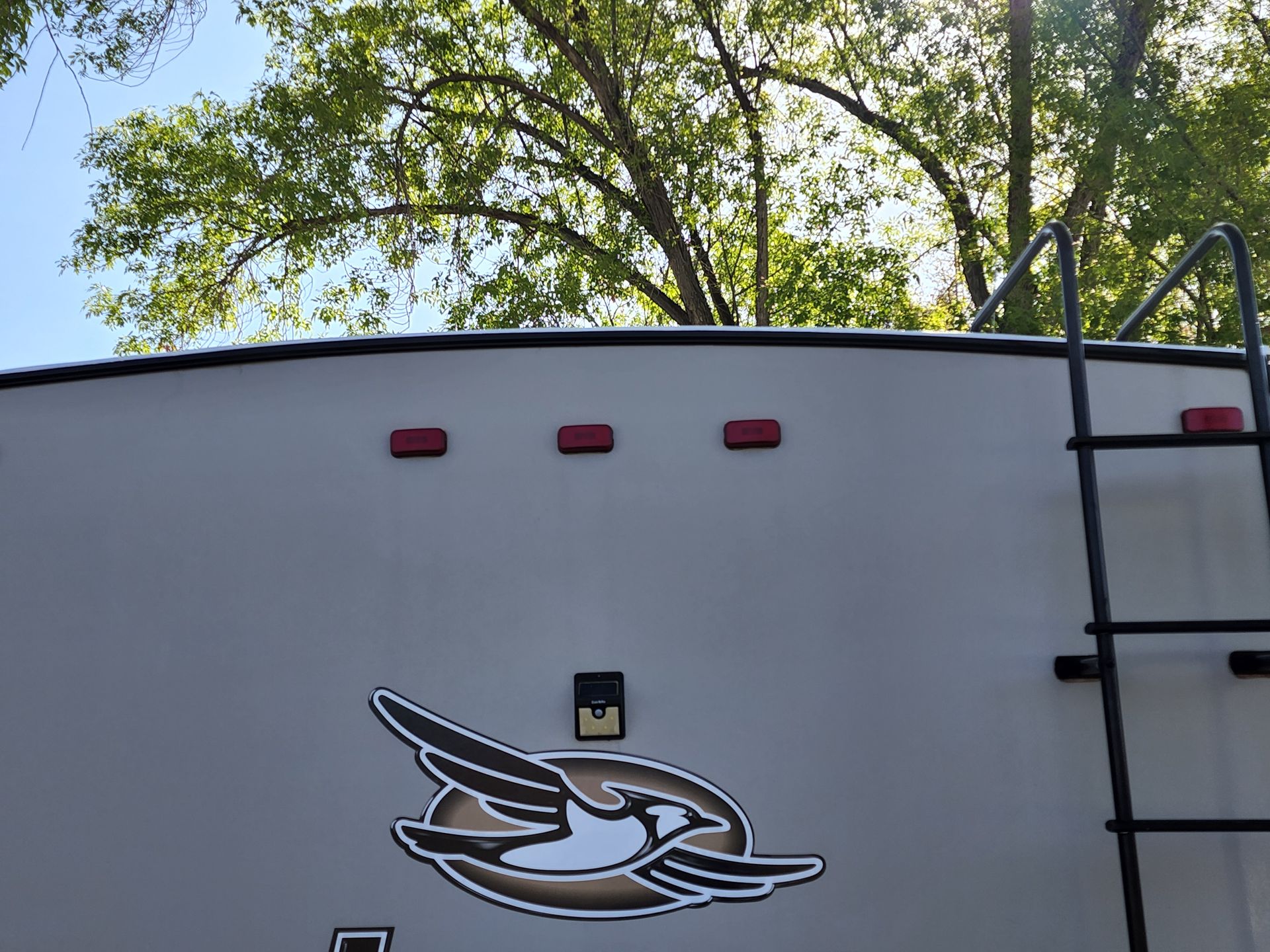 Tan RV rear with three red lights, logo, ladder, and trees in background.