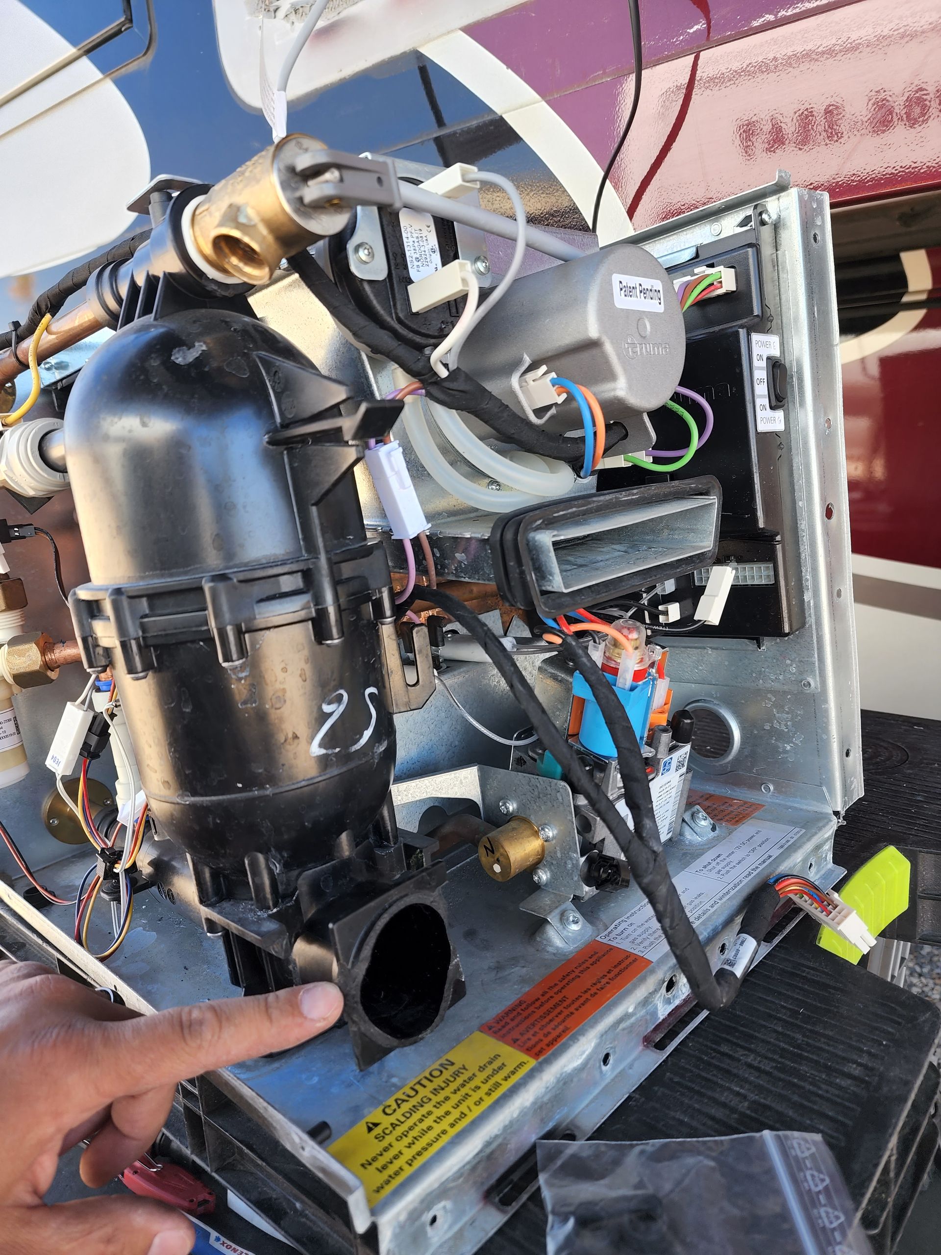 A hand points to a black component of a disassembled RV water heater. Wires and other parts are visible.
