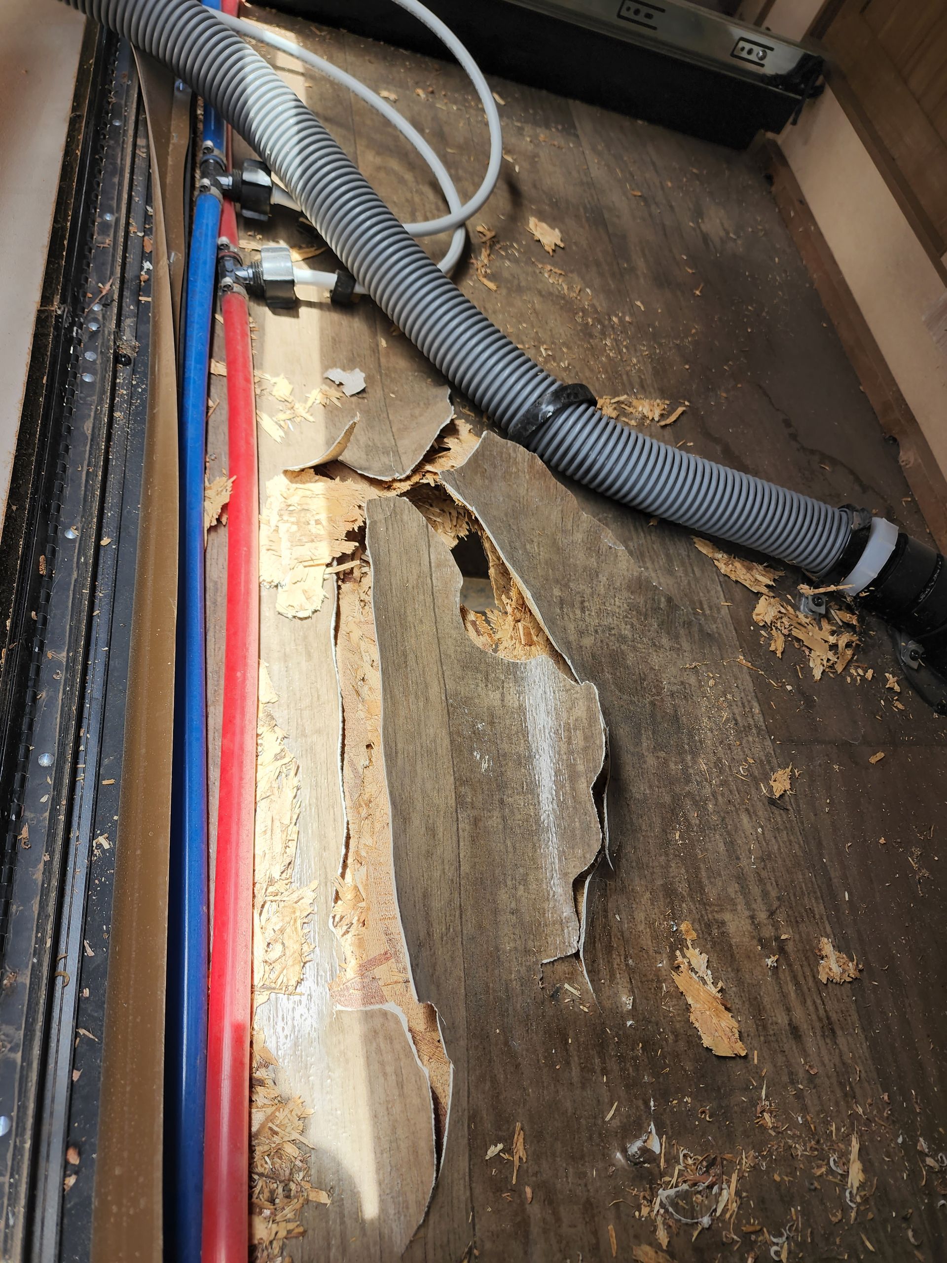 Damaged wooden RV flooring with water pipes and a corrugated hose visible.