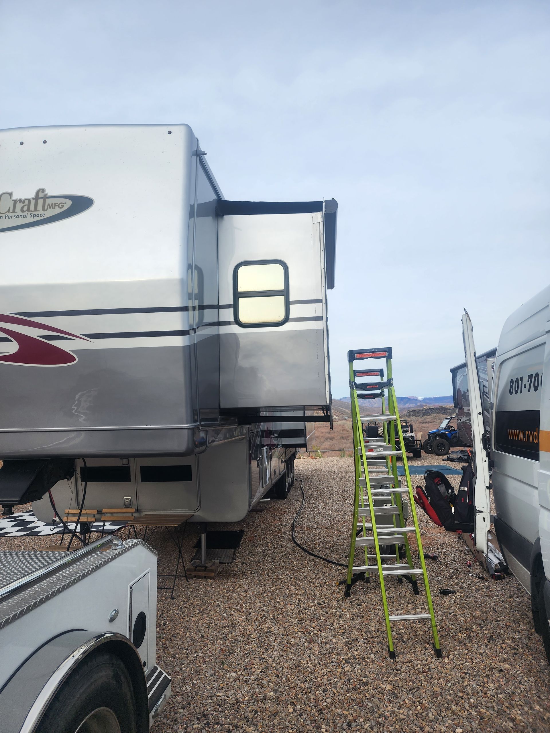 RV with extending slide-out, parked on gravel with a ladder and van in the scene.
