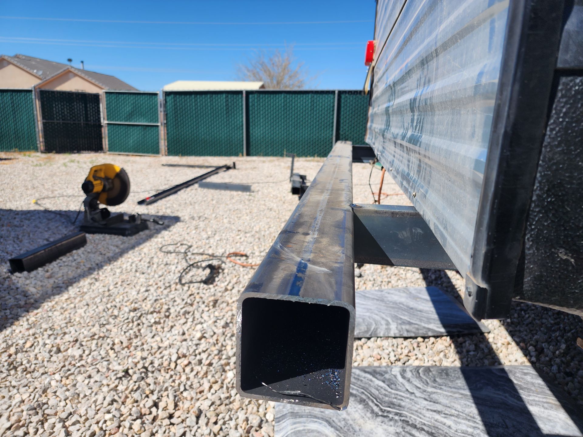 Metal square tube on gravel next to a saw and RV exterior, outdoors.