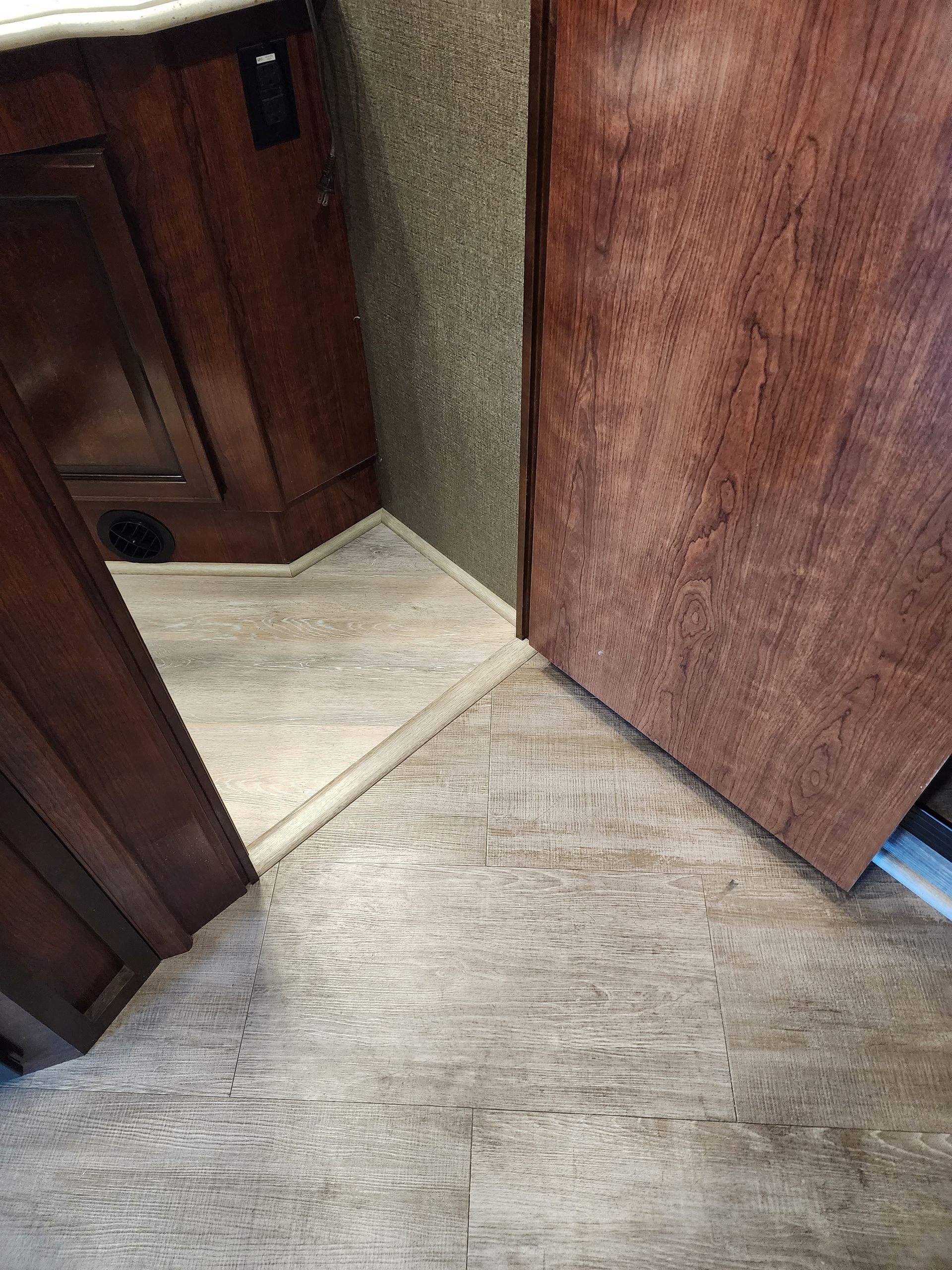 Interior view of a recreational vehicle. Flooring transitions to a textured, gray wall. Wooden door and trim.