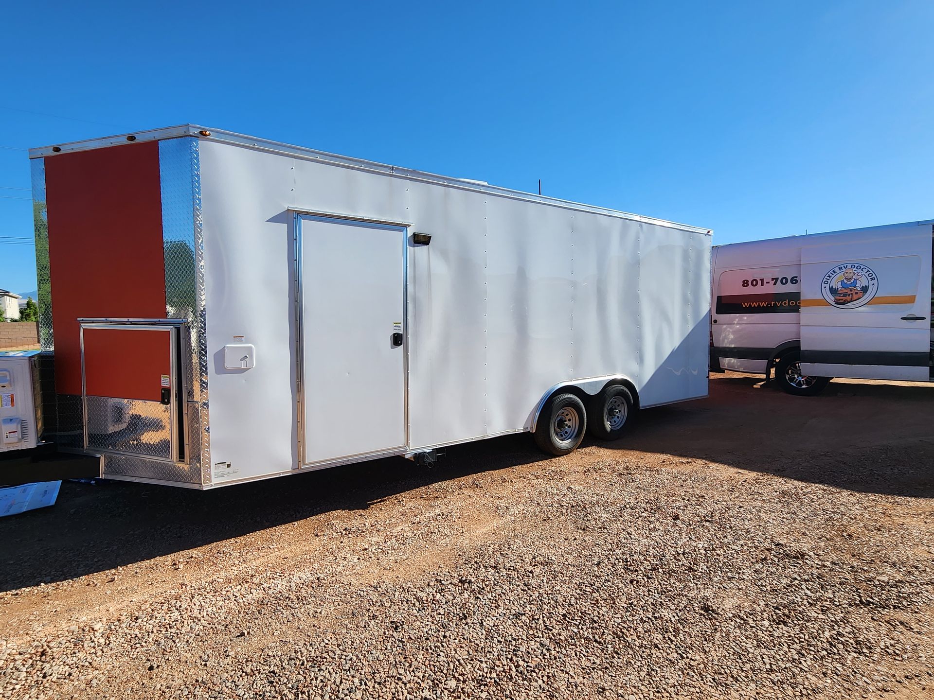 White enclosed trailer with an orange accent, parked on gravel, under a blue sky.