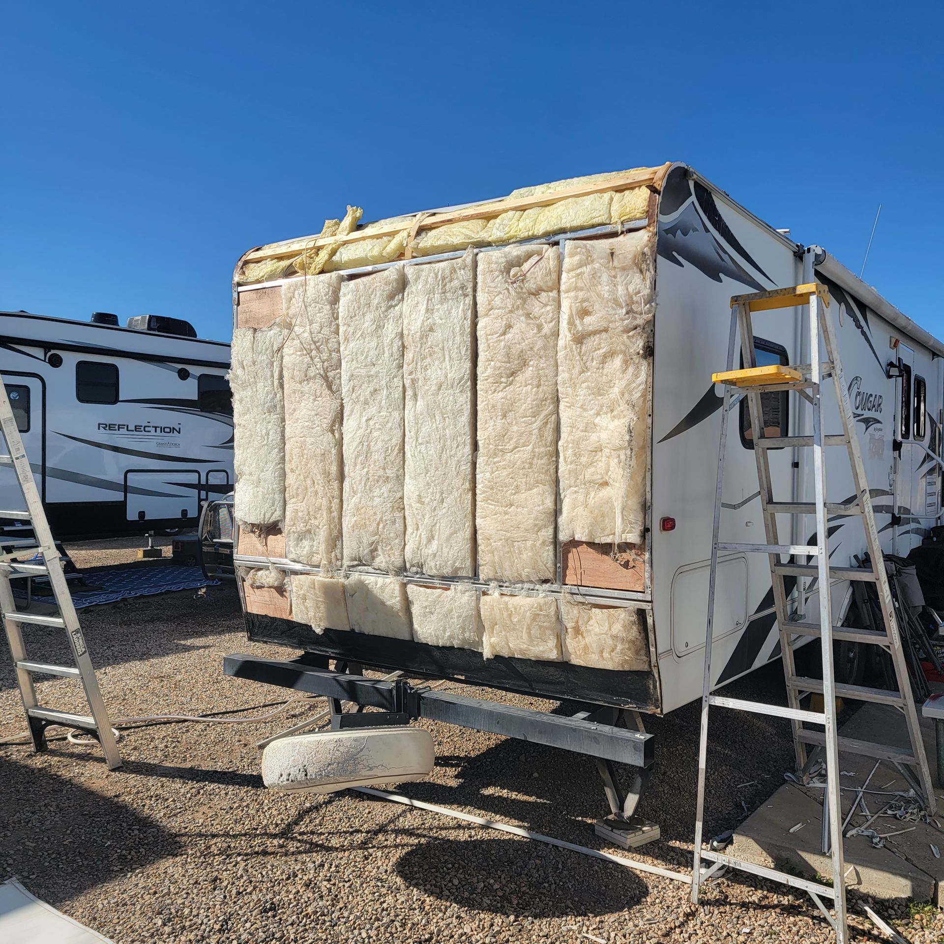 RV with exposed wall insulation being repaired outdoors on a sunny day; ladders nearby.