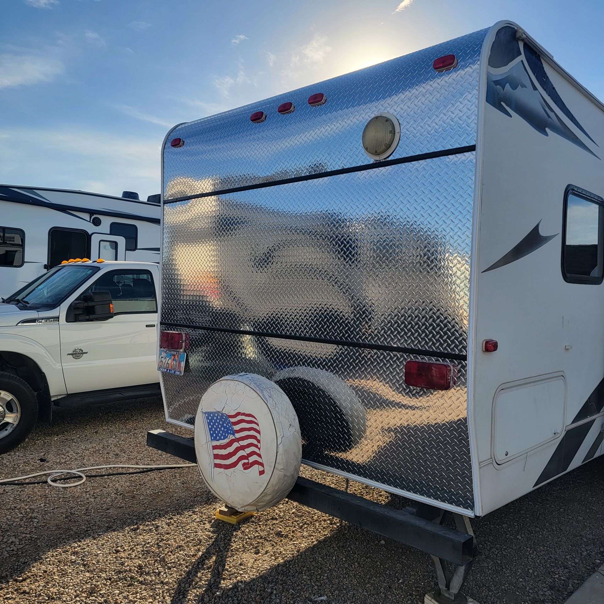 RV with diamond plate rear, white truck, tire cover with American flag, sunny setting.