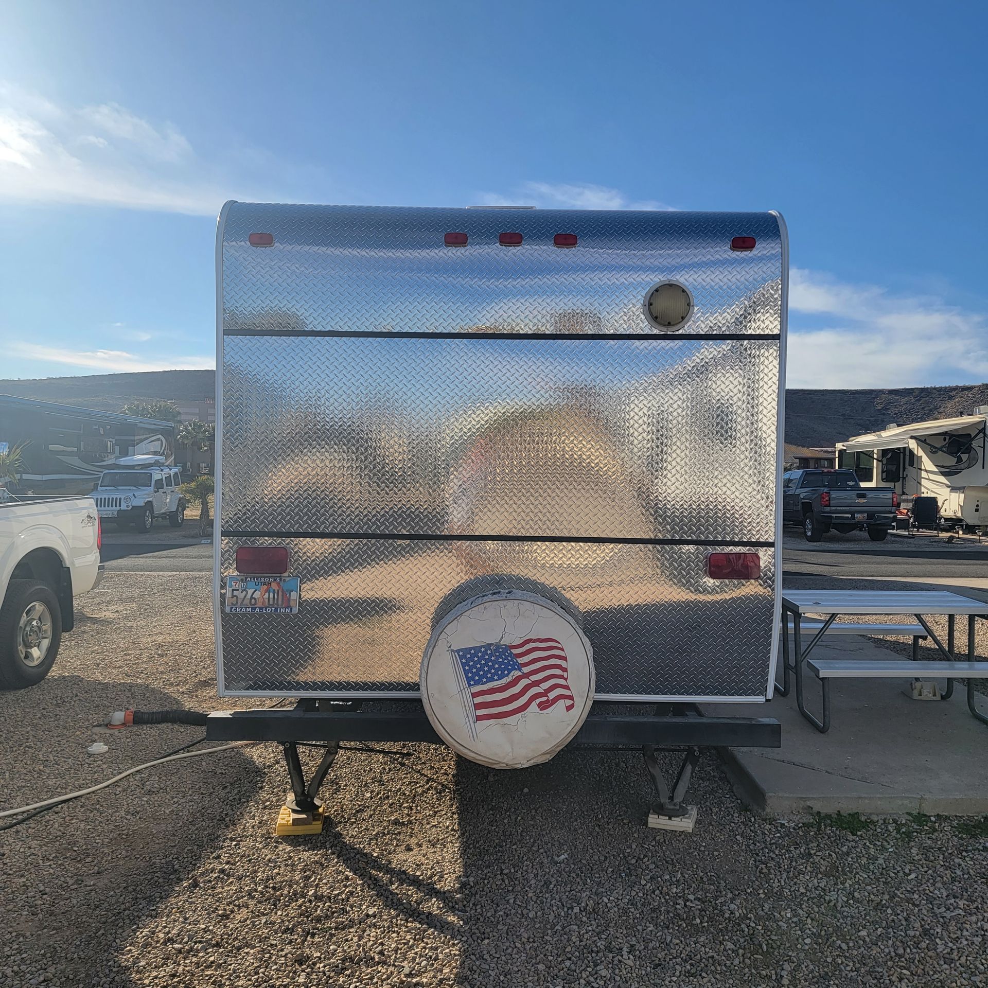Shiny aluminum travel trailer with American flag spare tire cover; parked at campsite.