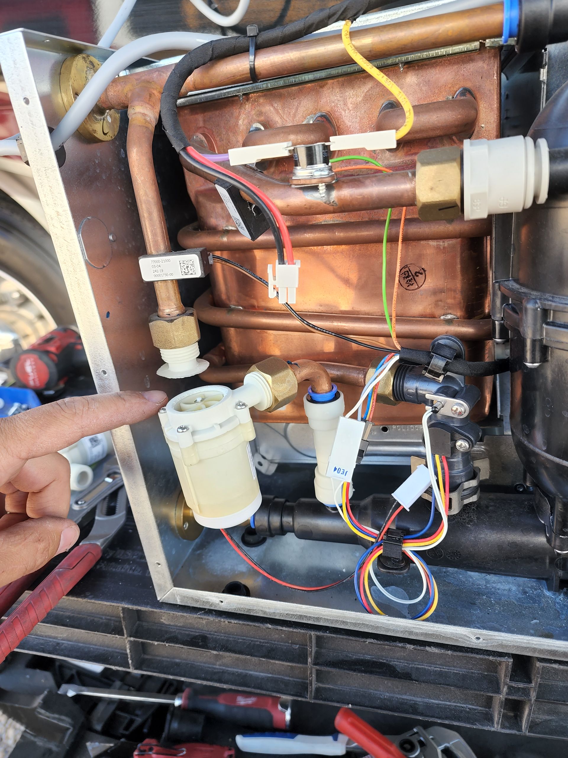 Hand pointing to a cylindrical water component within a machine's exposed interior, copper pipes visible.