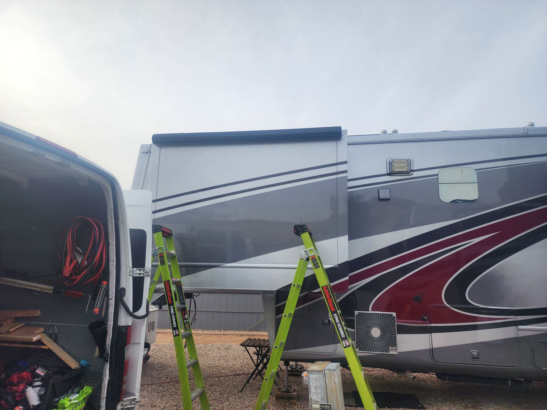 RV with extended awning. Two green ladders. Gray, red, and black exterior paint. Cloudy sky.