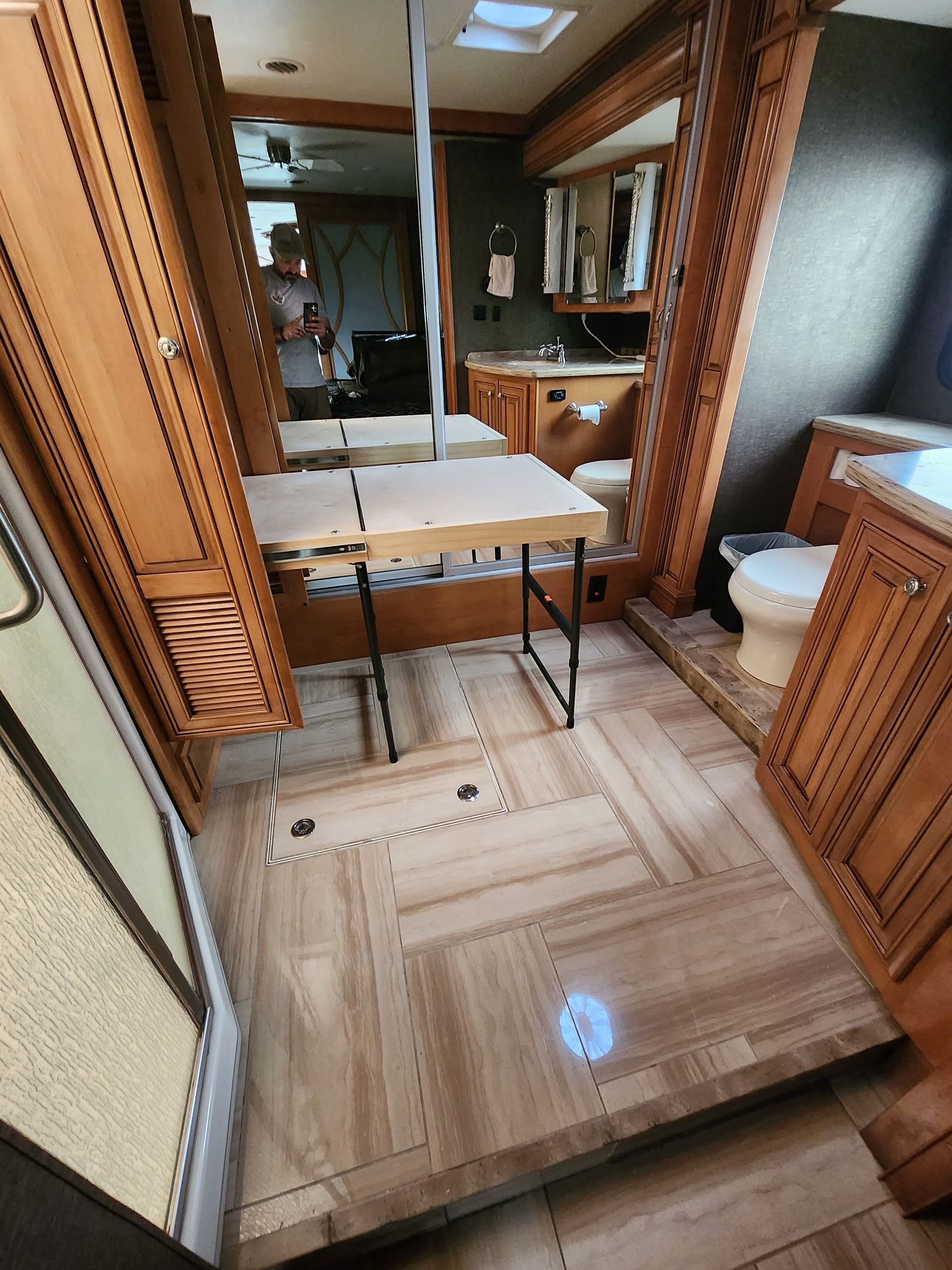 Interior of a recreational vehicle. Wooden floors, cabinets and walls. A fold-out table is in the center, and the bathroom is in the background.
