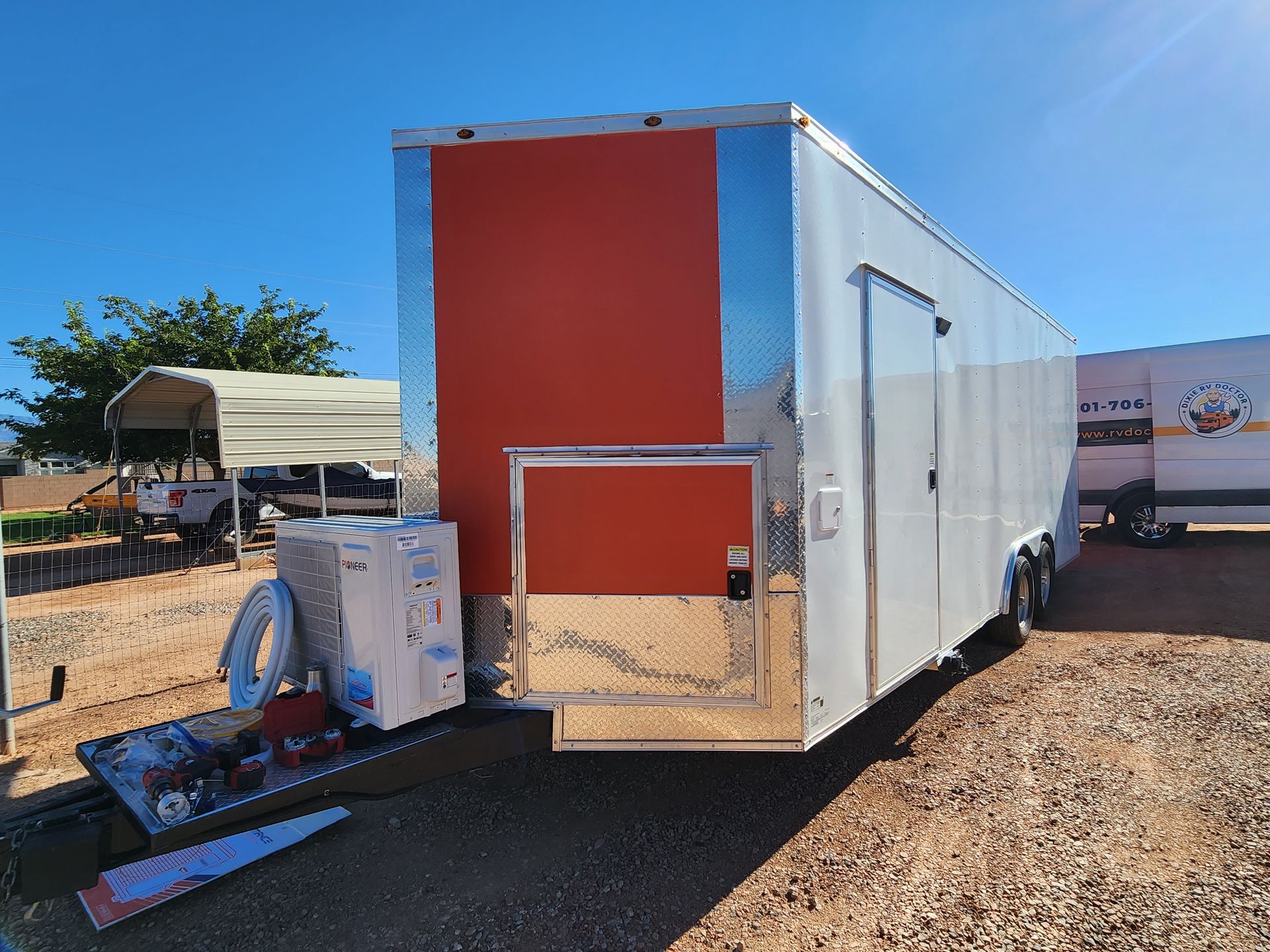 Enclosed trailer with silver siding and an orange accent panel, parked outdoors on gravel.