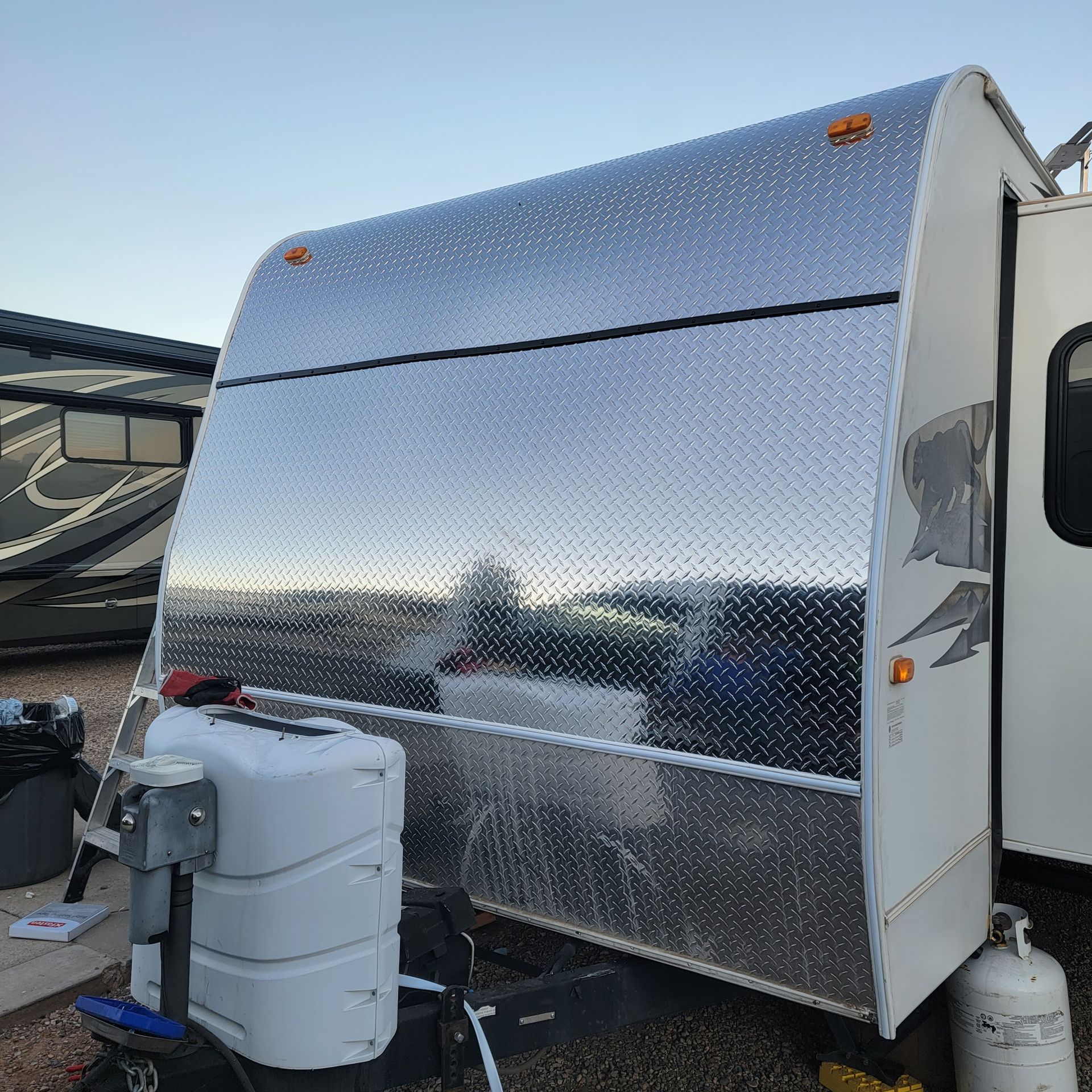 Shiny, diamond-plated travel trailer parked outdoors. Propane tank and hitch visible in front. Open door on the right.