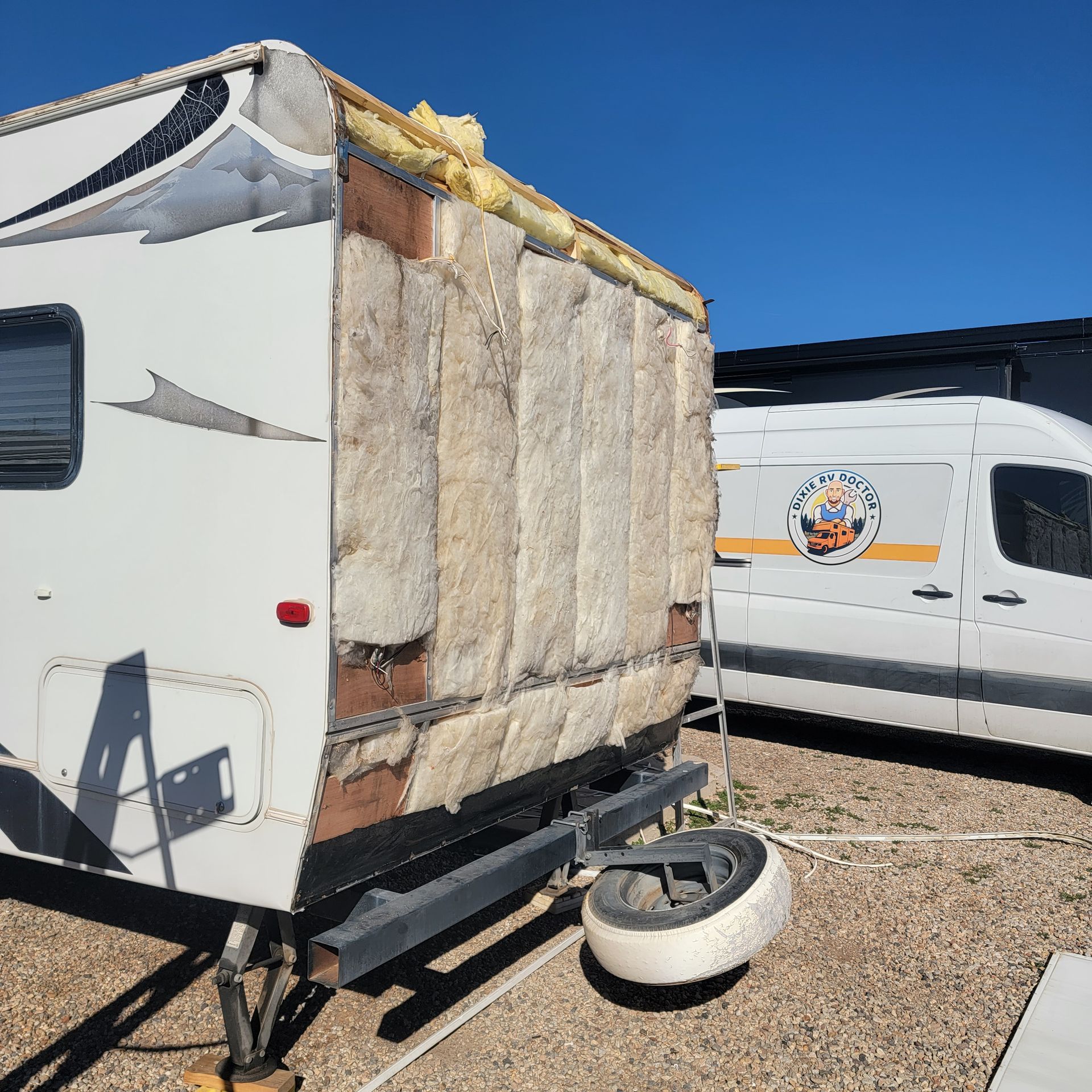 RV with side panel removed, exposing insulation. White van parked nearby.