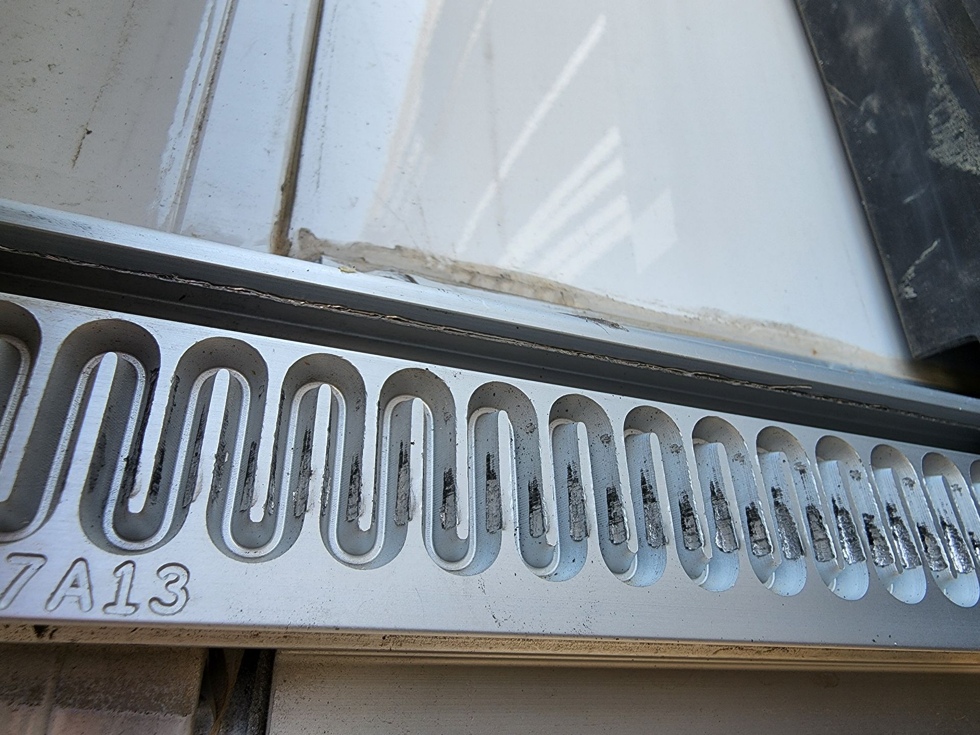 Accordion-like expansion joint, silver metal, installed between a white surface and a dark corner.