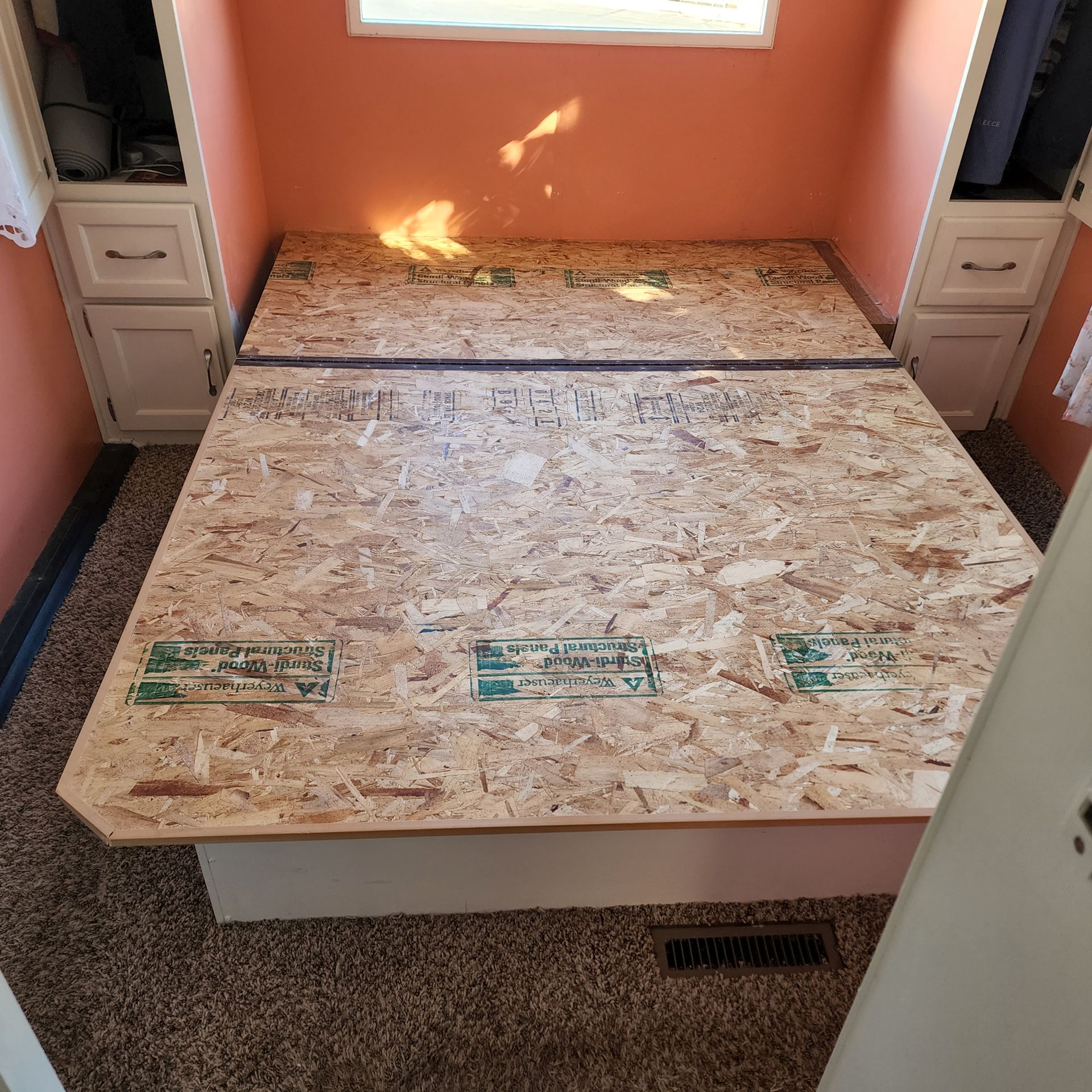 OSB bed frame in a peach-colored room, flanked by white cabinets. Carpeting on the floor.