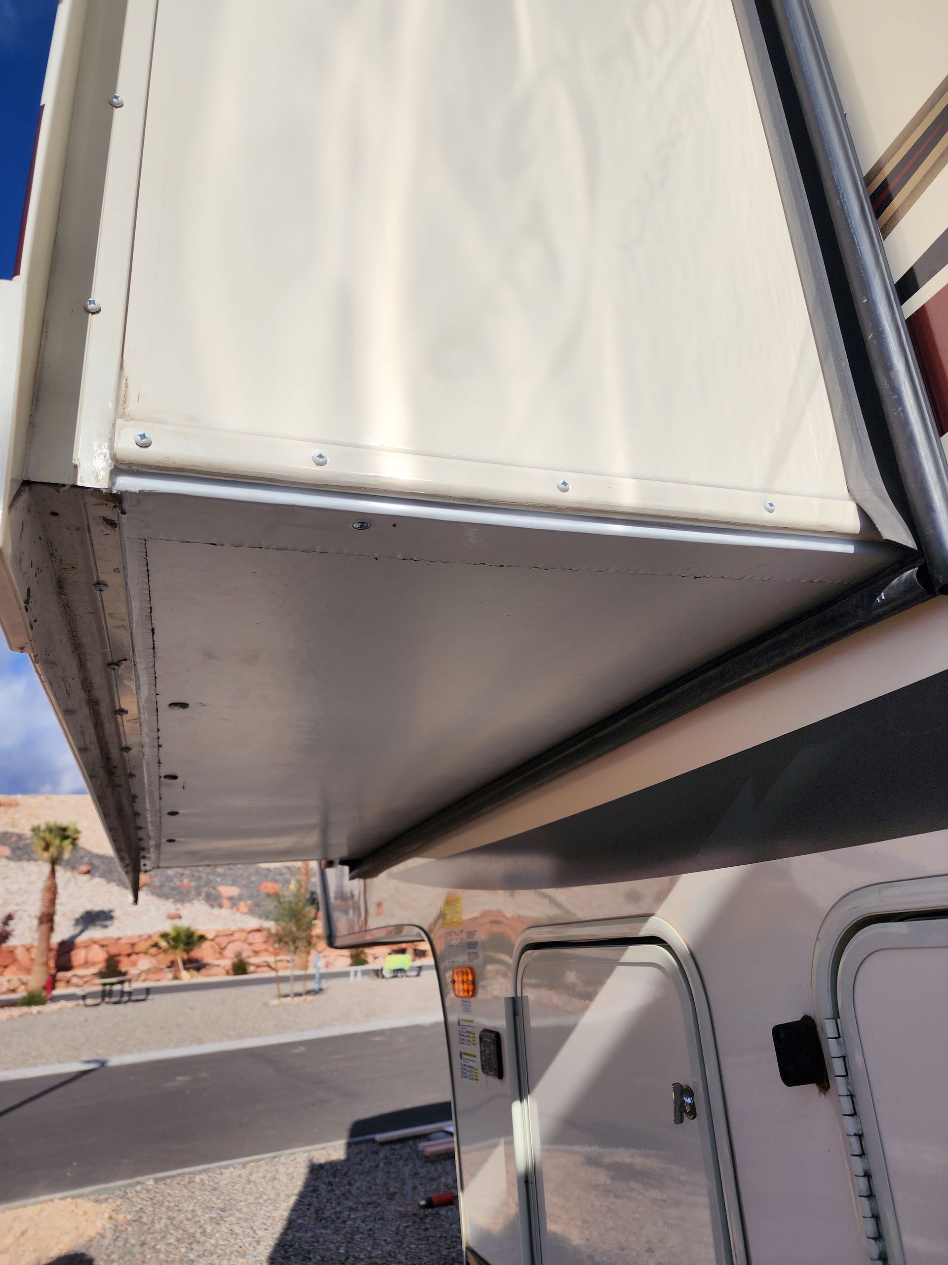 A close-up of the side of a white RV. The top section is a raised overhang with a white awning.