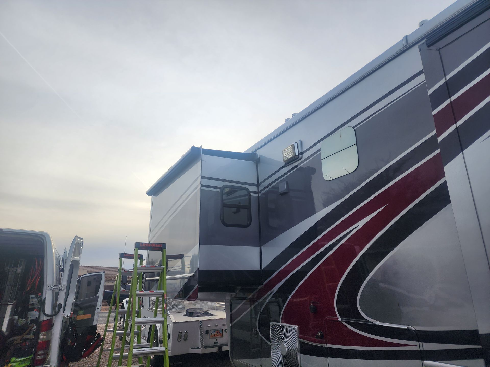 RV exterior with extending slide-out; silver, maroon and red accents against cloudy sky. Ladder and equipment present.