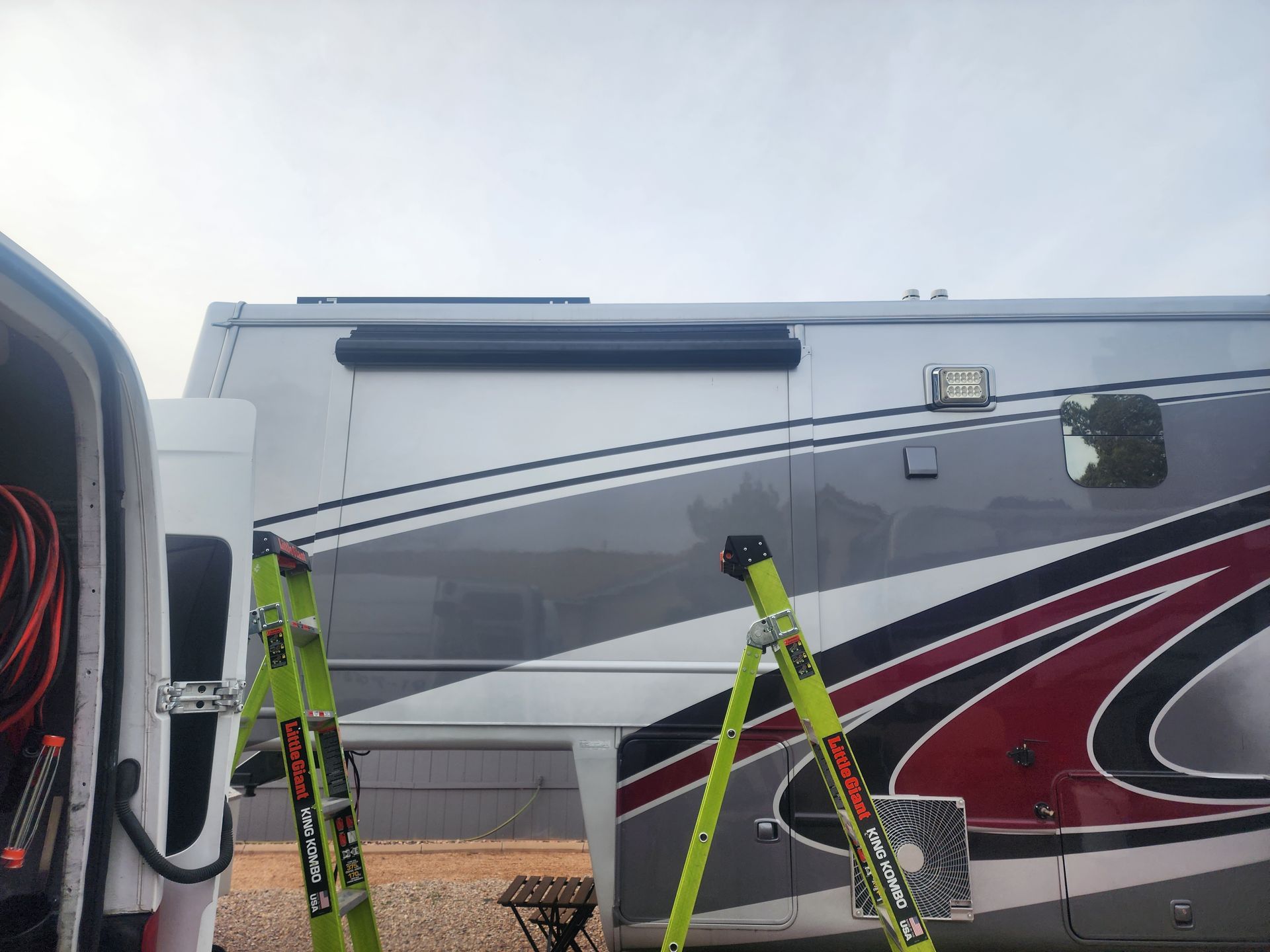 A motorhome with a black awning extended, with ladders nearby, and a vehicle is to the left.