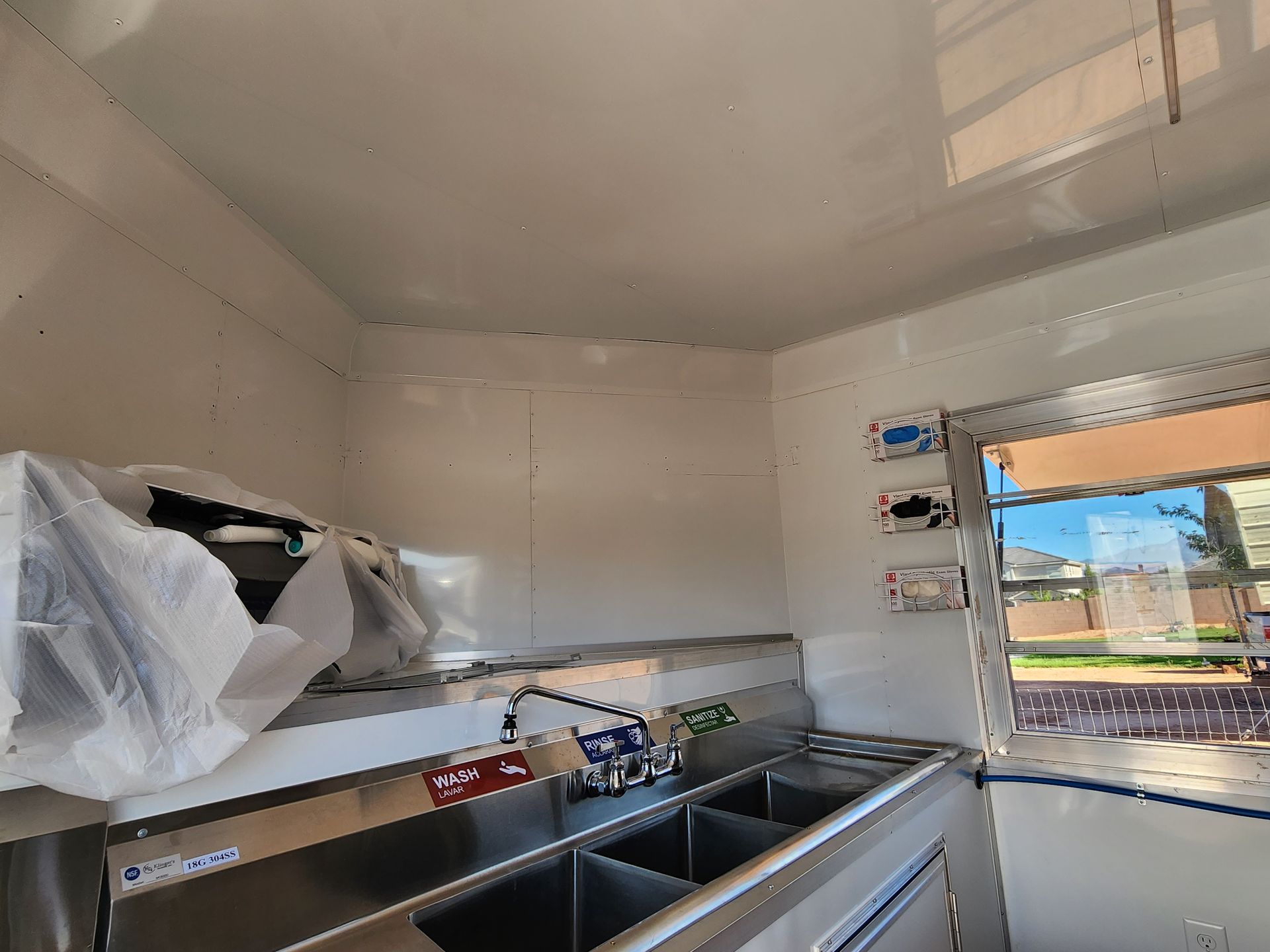 Interior of a food truck. Stainless steel sinks and counter, white walls, and a window reveal an outside view.