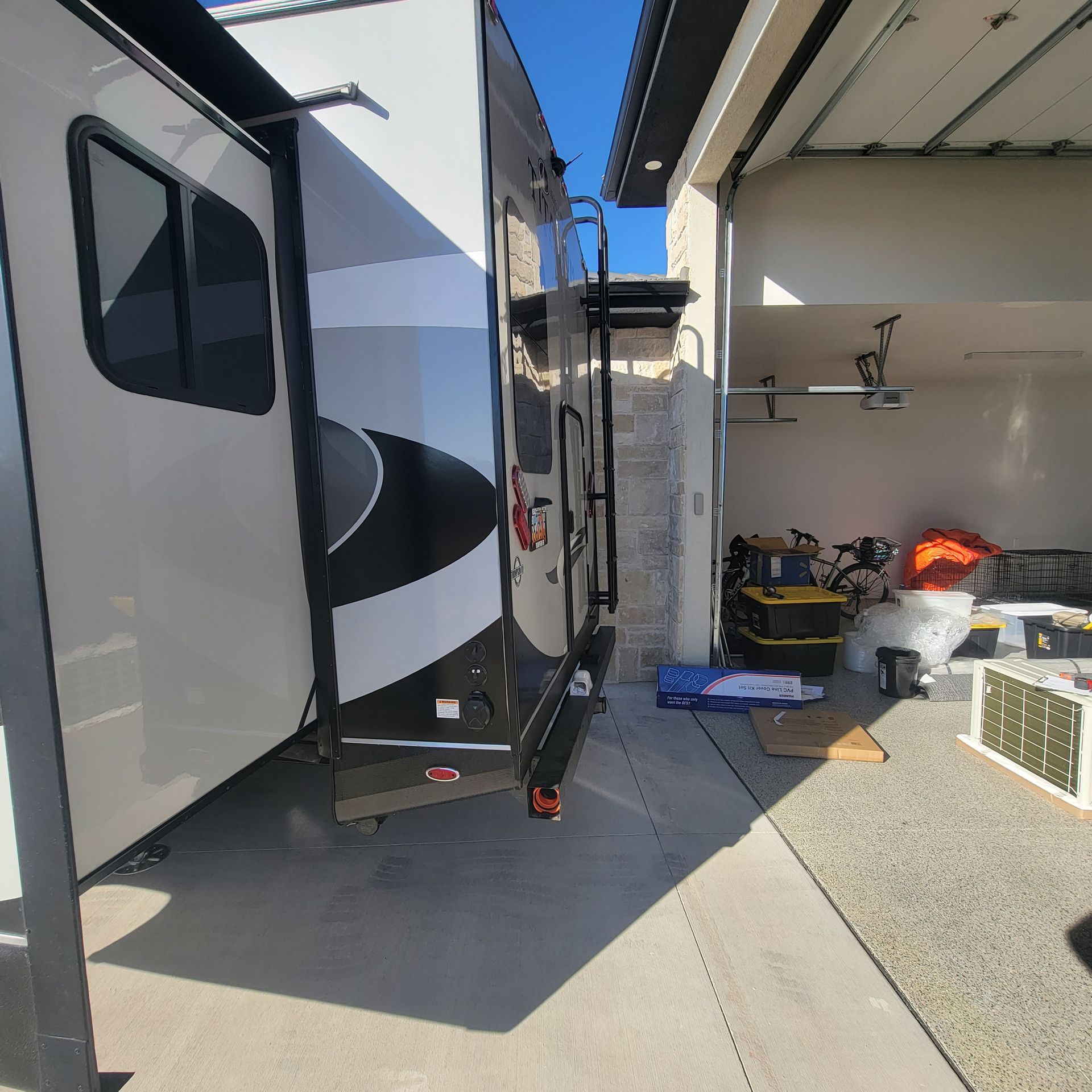 RV parked next to a garage. Narrow space between the vehicles. Bright sunlight.